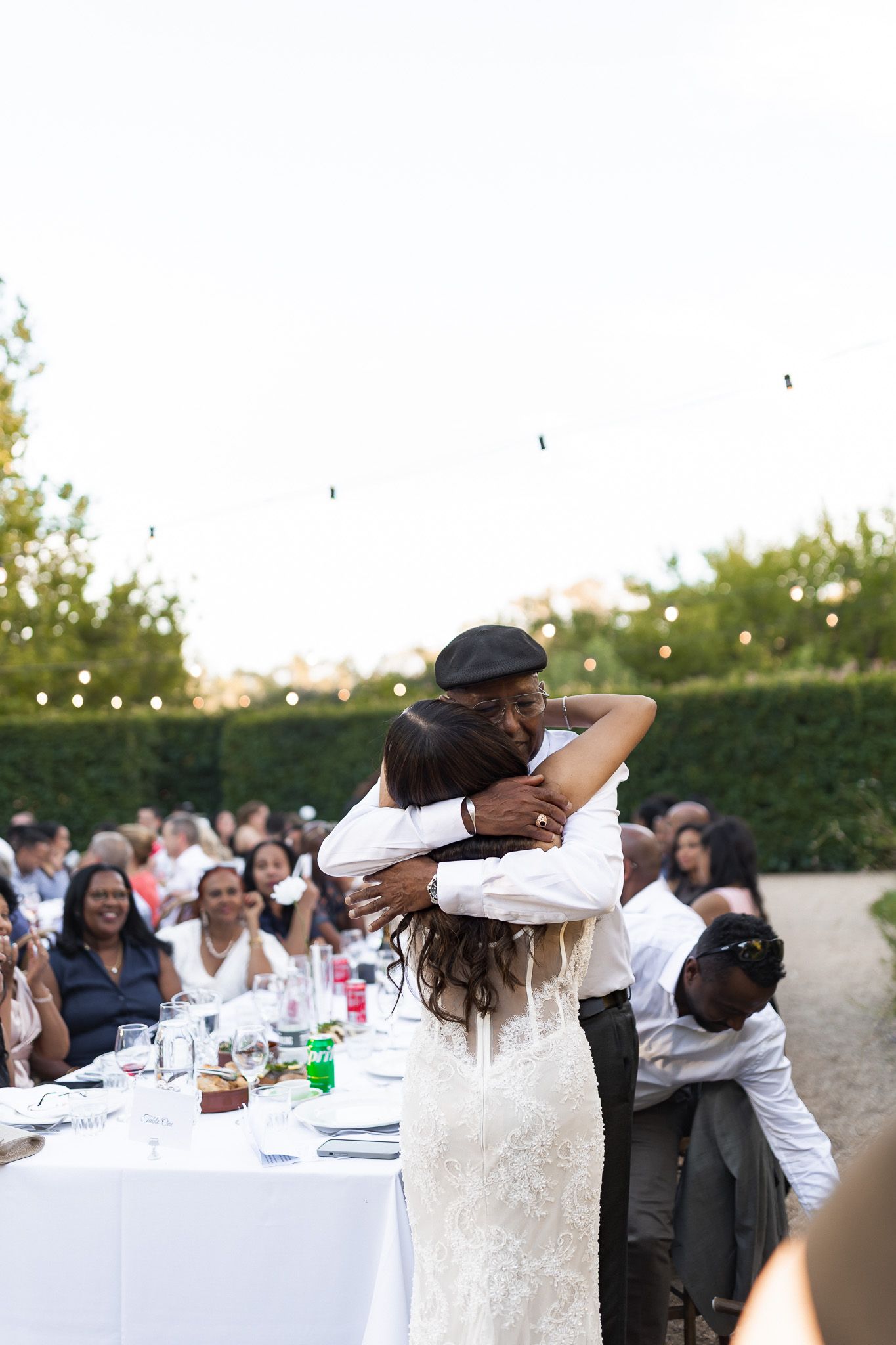 Bride in lace gown sharing a warm embrace with an older guest at an outdoor wedding reception.
