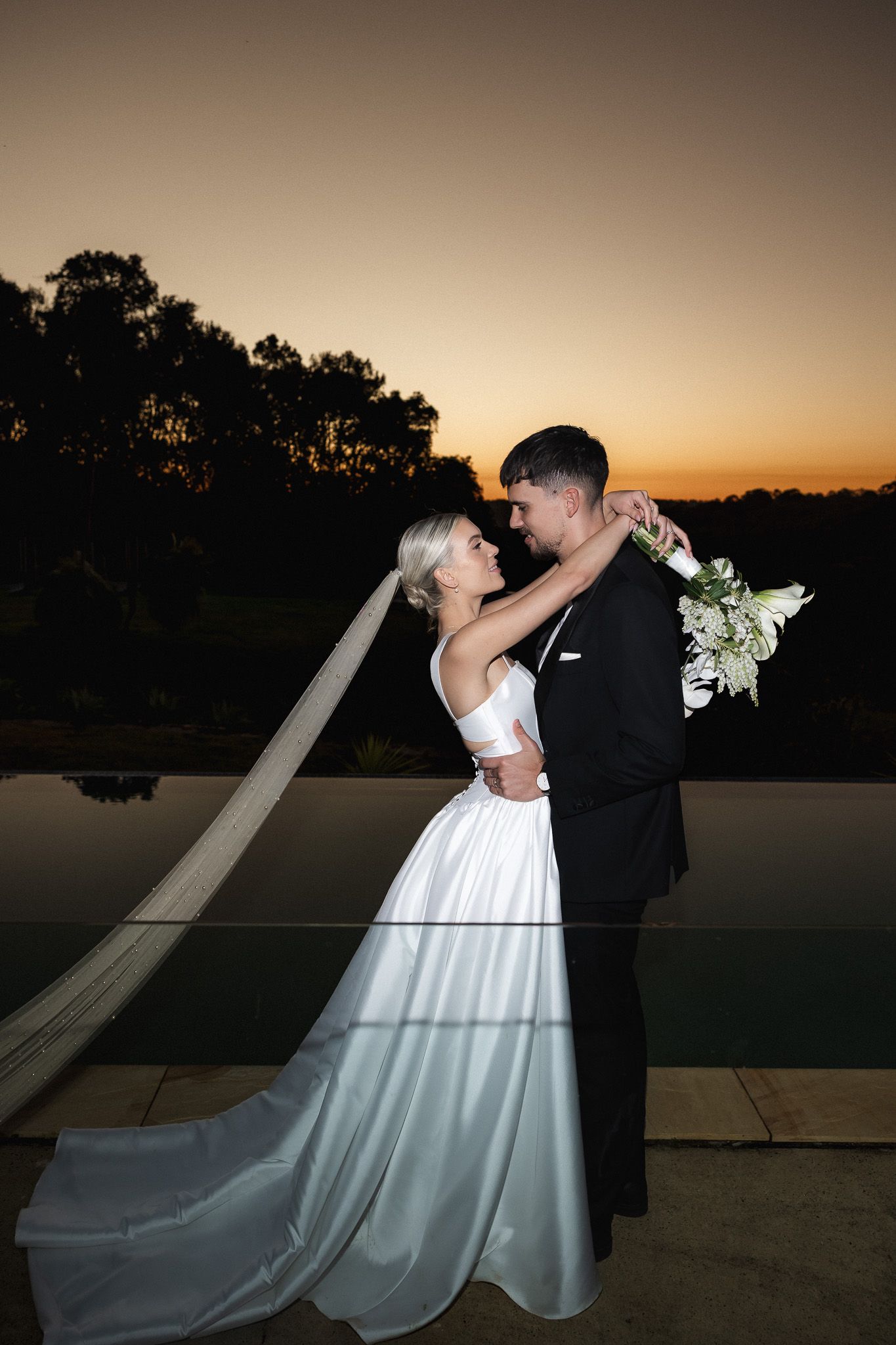 Bride and groom share romantic embrace at sunset beside infinity pool, bride holding white floral bouquet.