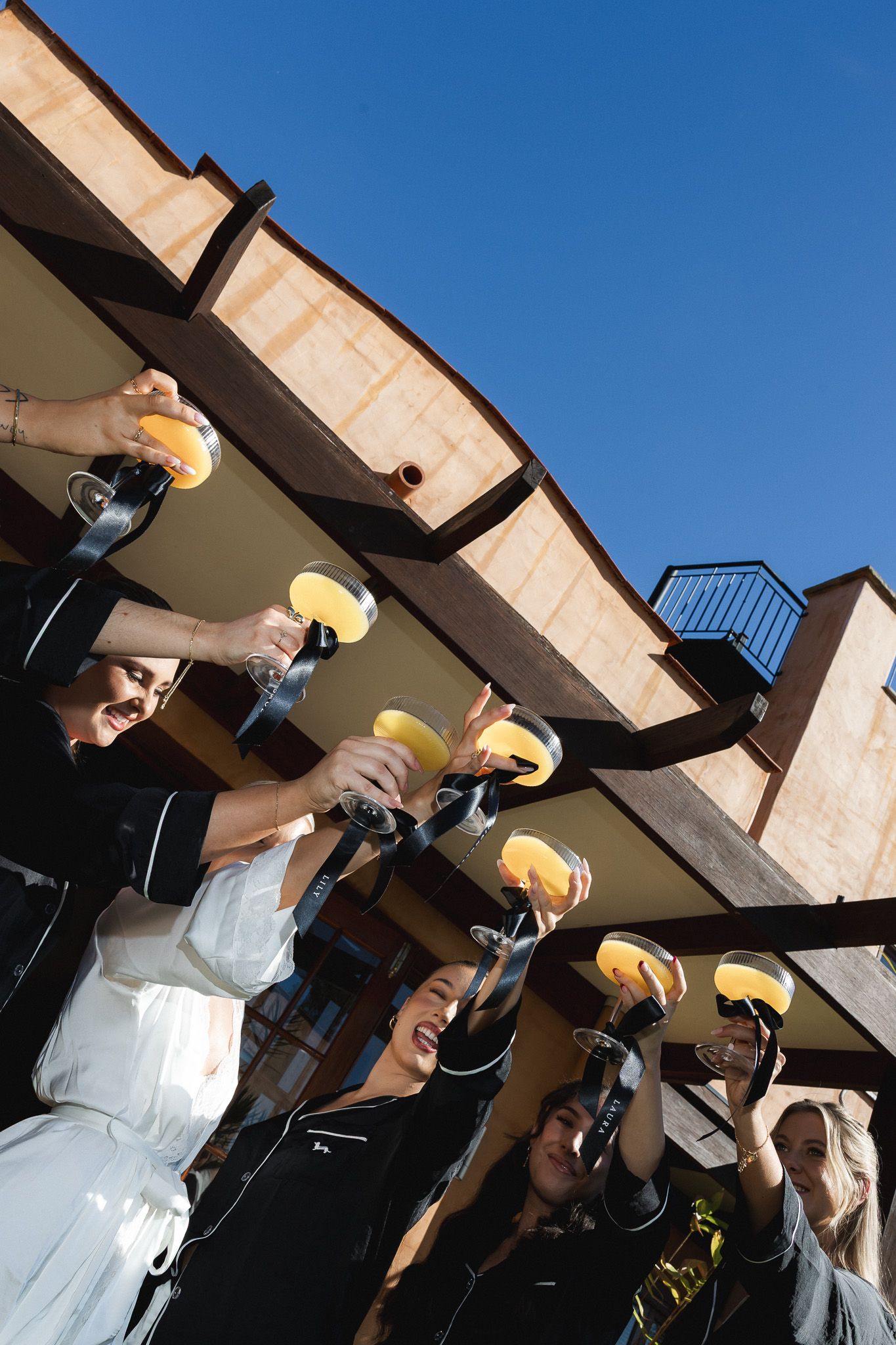 Bridesmaids in black robes raising yellow cocktails in celebration under blue sky on balcony.