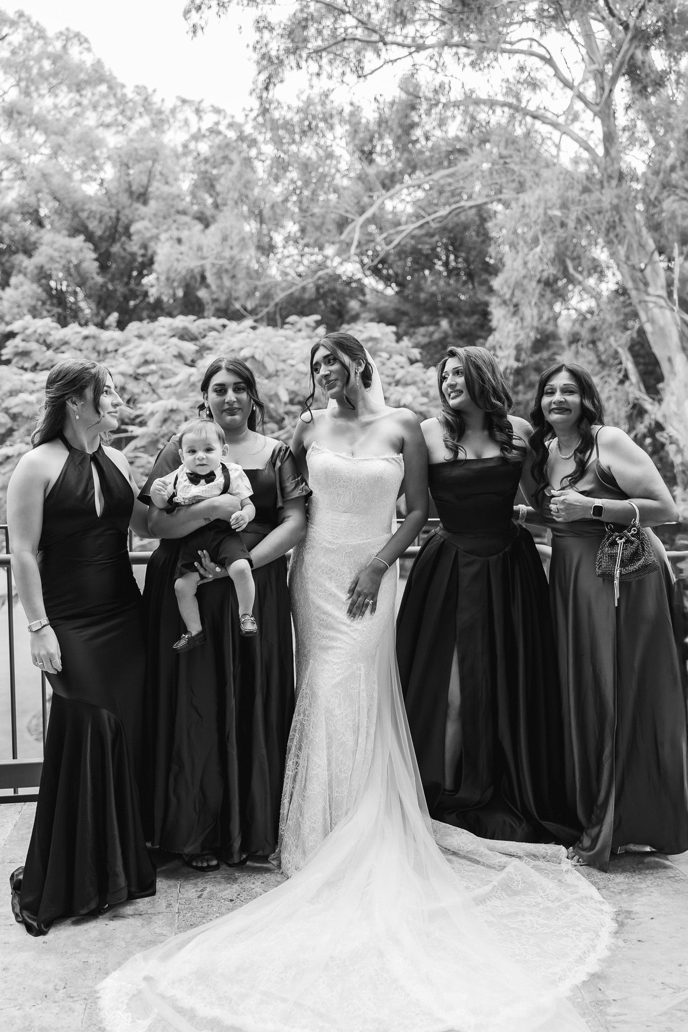 Black and white photo of bride in lace gown posing with bridesmaids in black dresses outdoors.