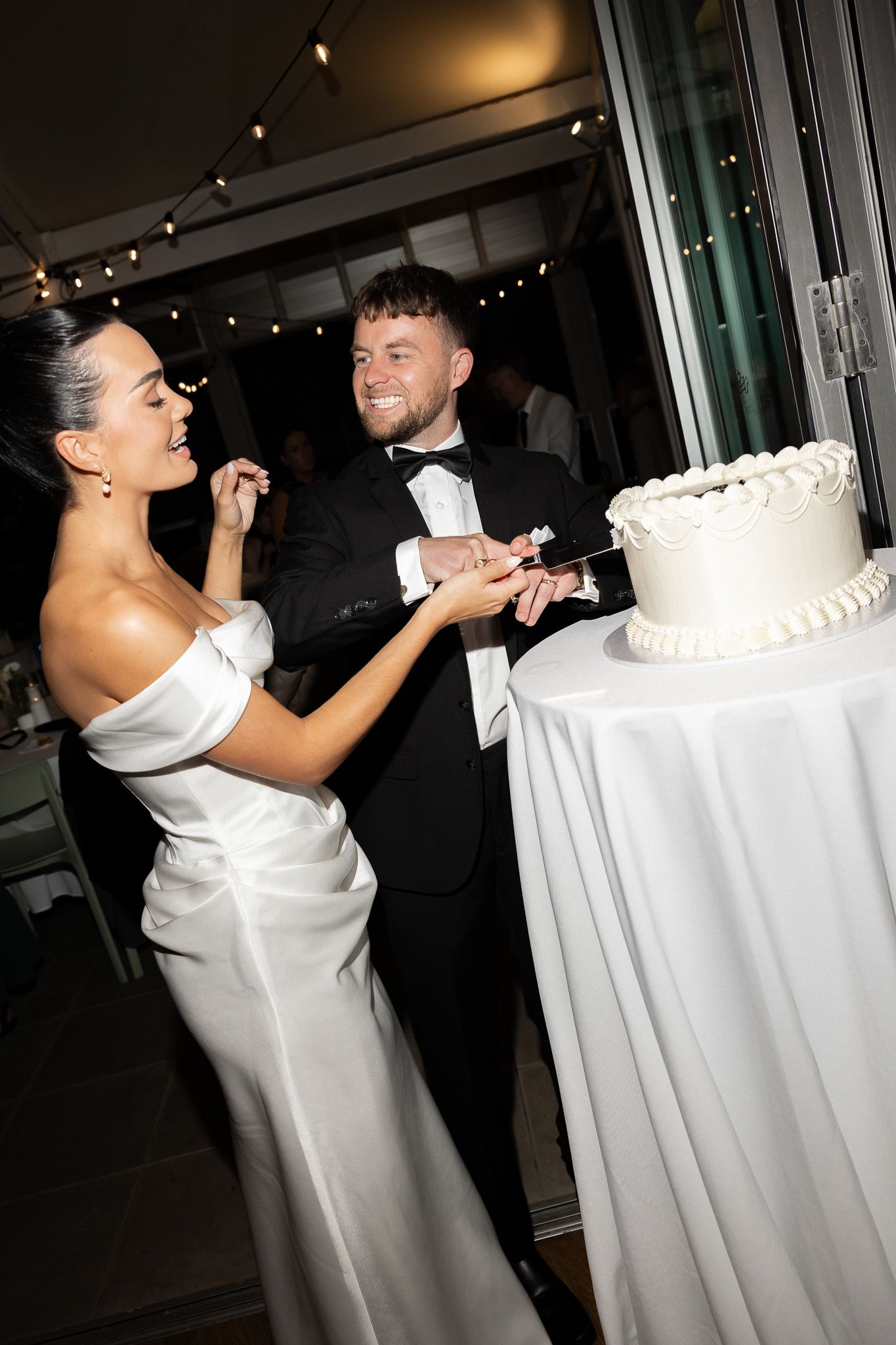 Bride and groom in formal attire smile while cutting white wedding cake at reception.