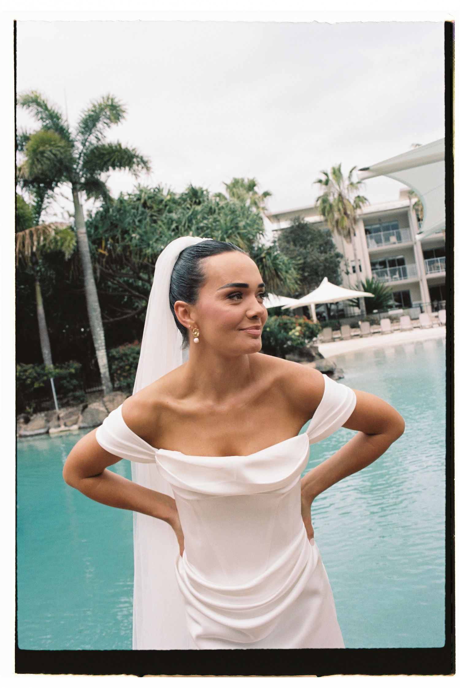 Bride in off-shoulder white gown and veil poses by tropical pool with palm trees and resort building.