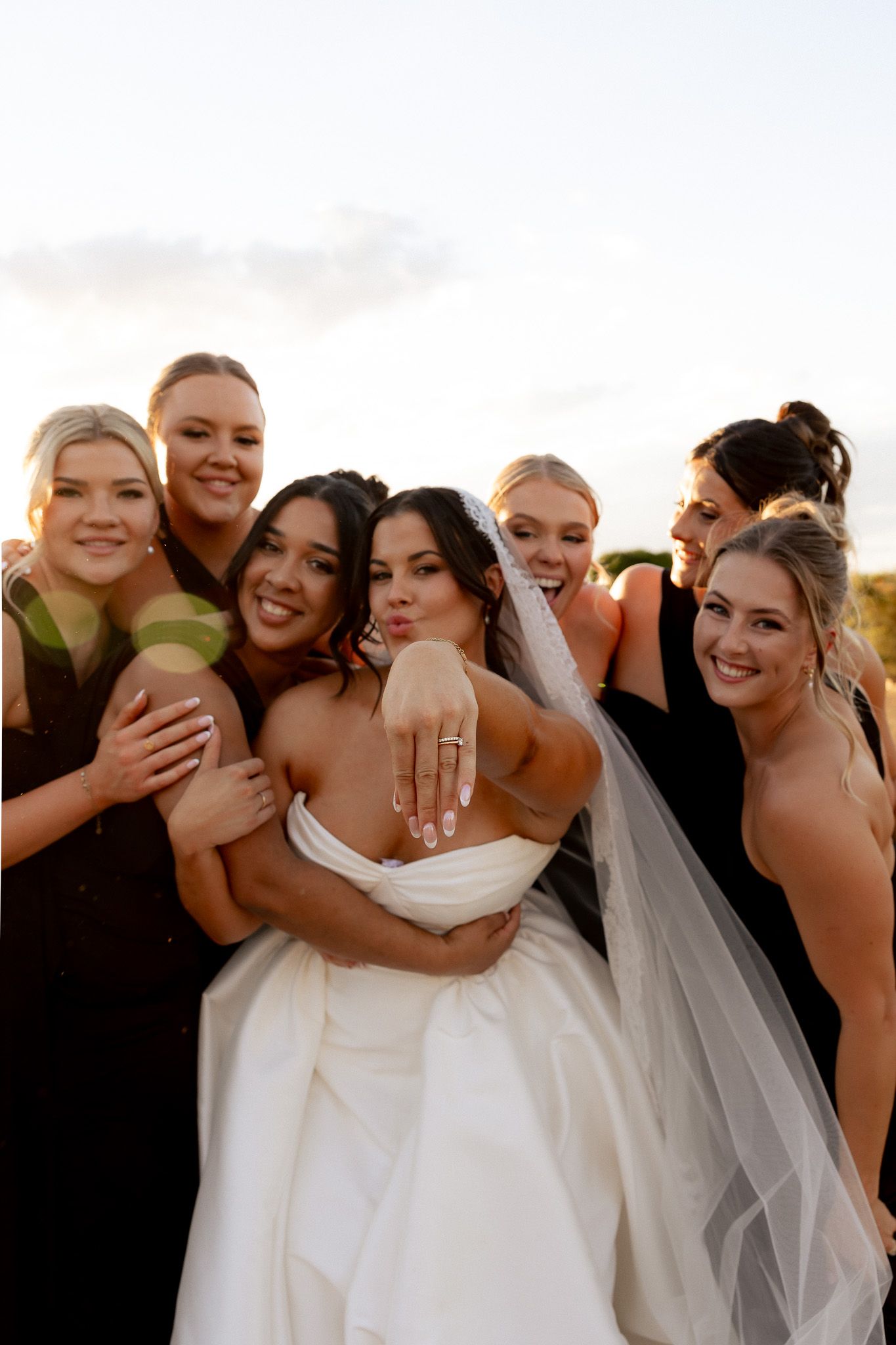 A smiling bride in a white gown and veil poses with five bridesmaids in black dresses at golden hour.