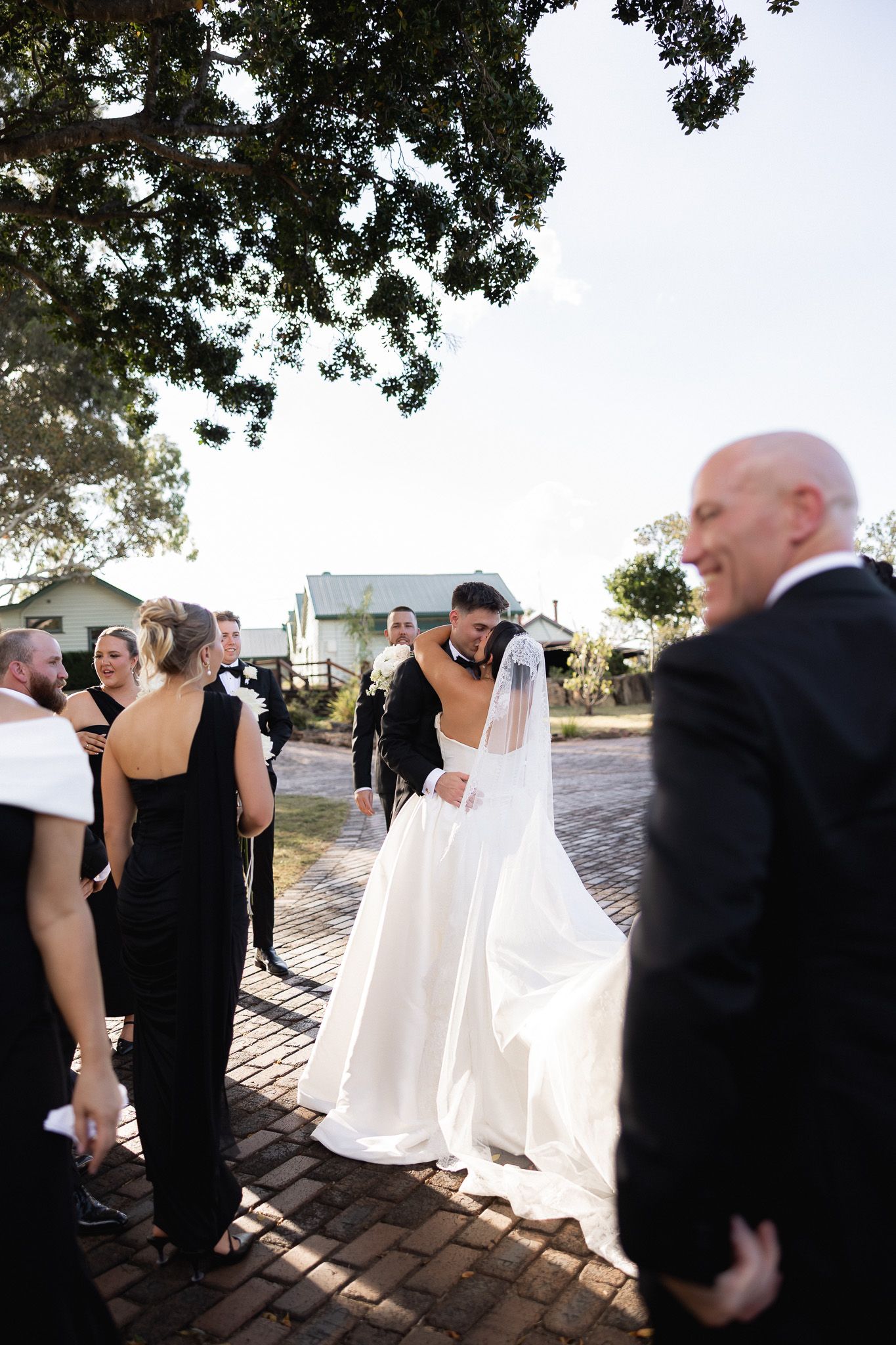 Groom embracing bride in white wedding gown surrounded by wedding party on cobblestone path.