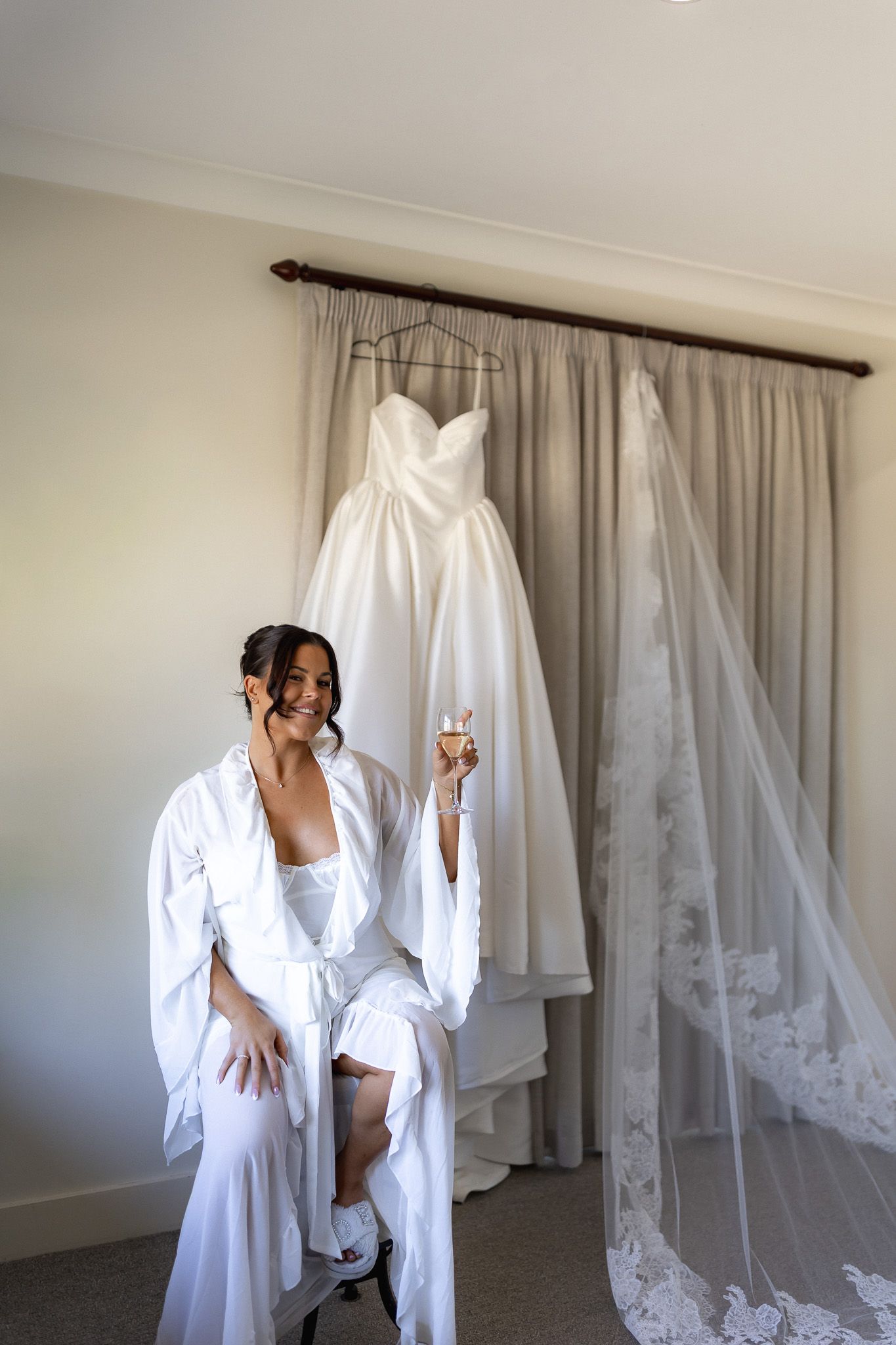 Bride in white robe holding champagne glass seated before her elegant wedding dress and lace veil hanging on curtain rod.