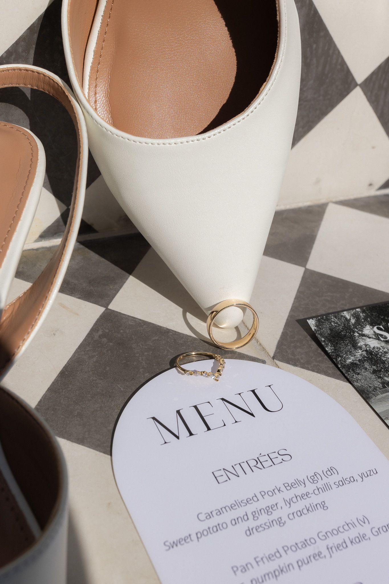 White bridal heels with gold wedding rings resting on an elegant wedding menu card on a checkered surface.