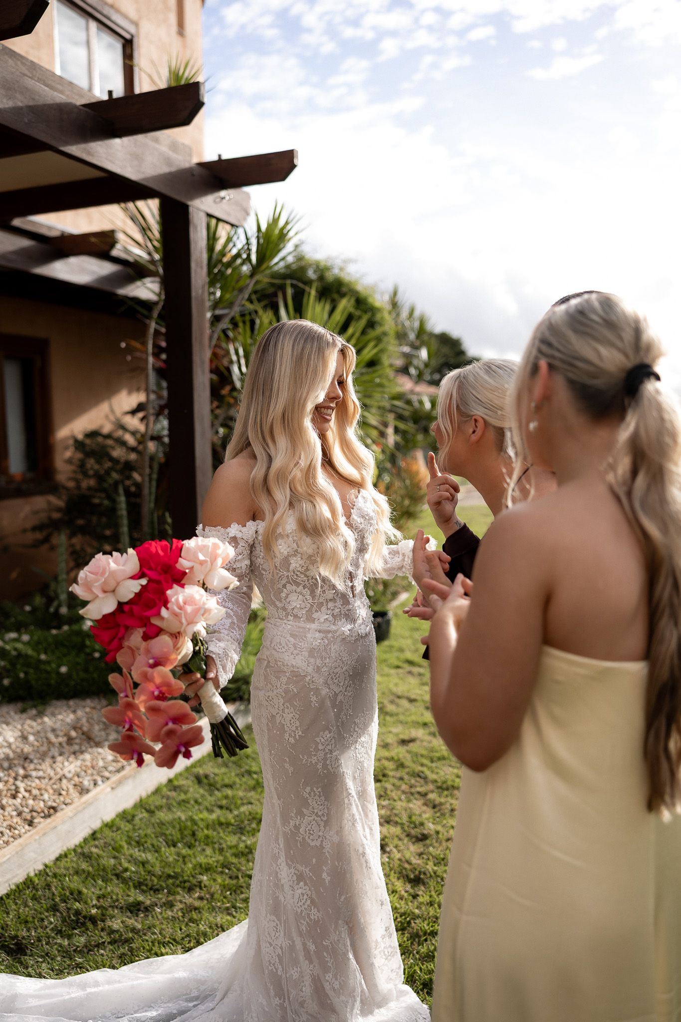 Bride in lace gown holding pink bouquet with bridesmaids in yellow dresses celebrating outdoors at a destination wedding.