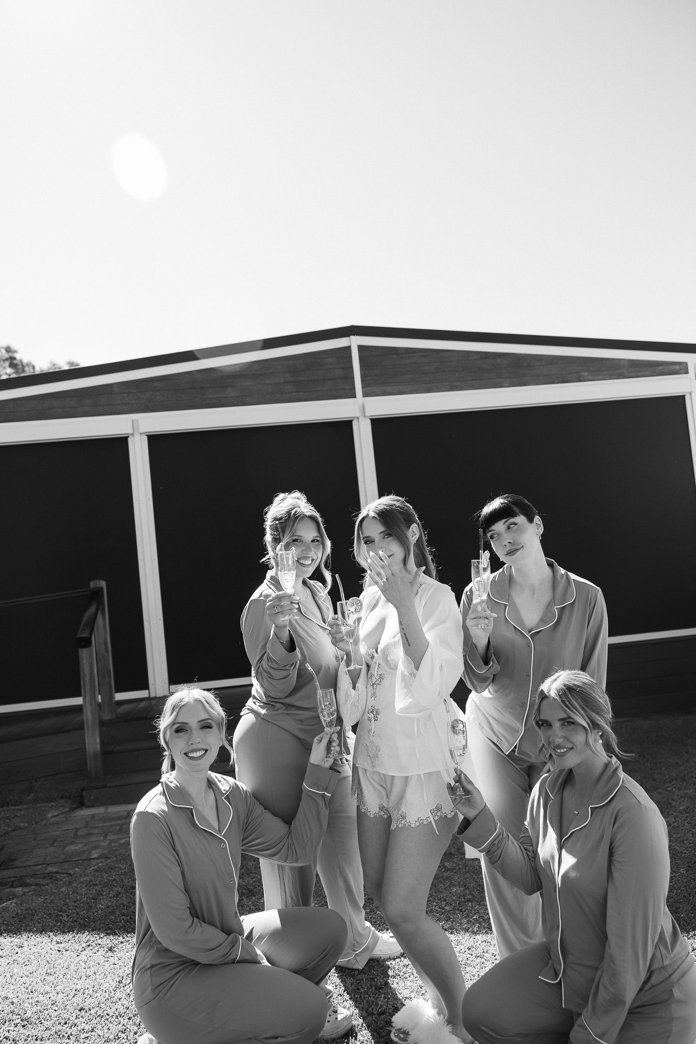 Black and white photo of a bride and bridesmaids in matching pajamas toasting with champagne glasses outdoors.