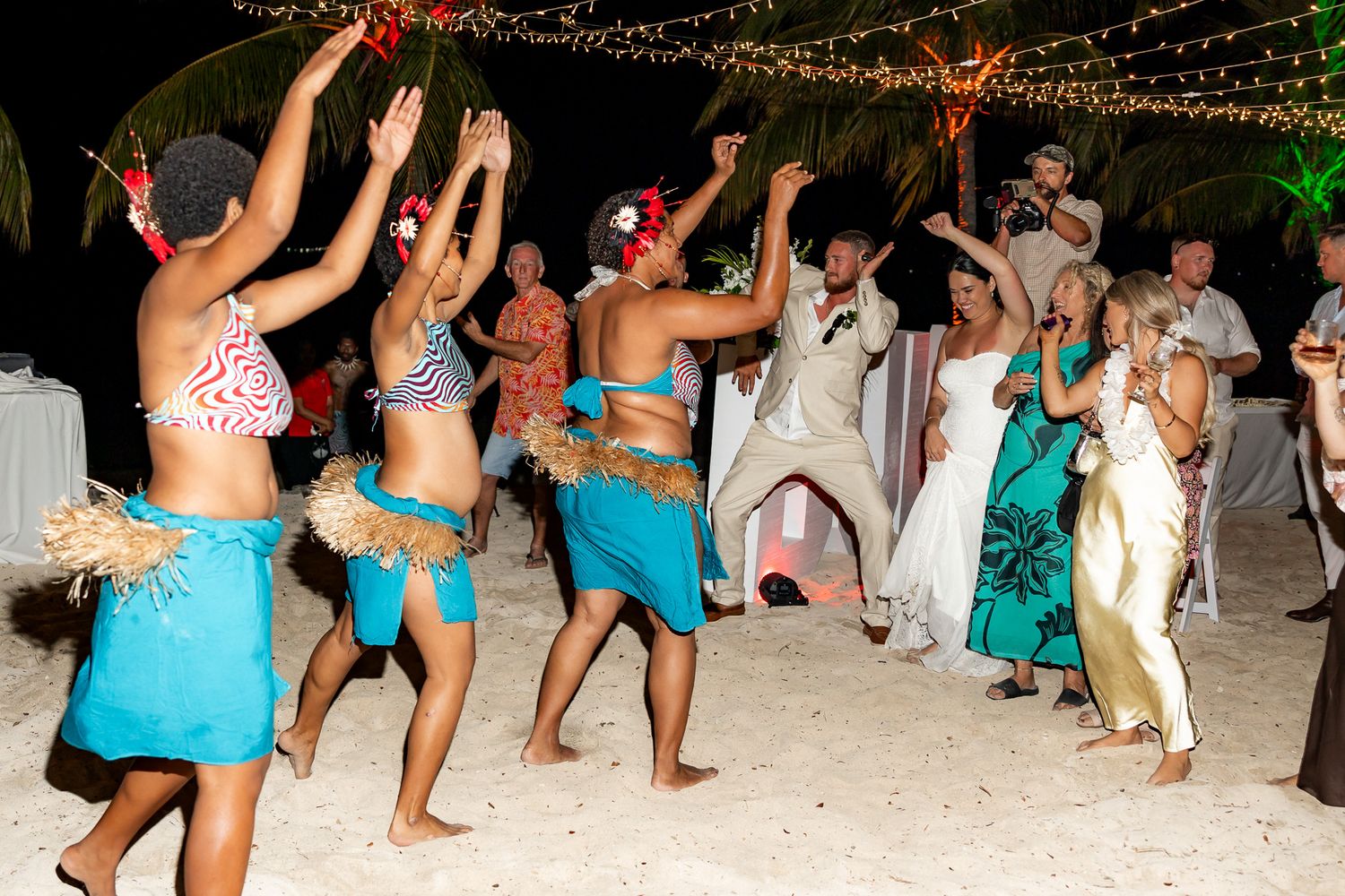 Wedding guests and couple dance with traditional Polynesian performers on sandy beach at night.