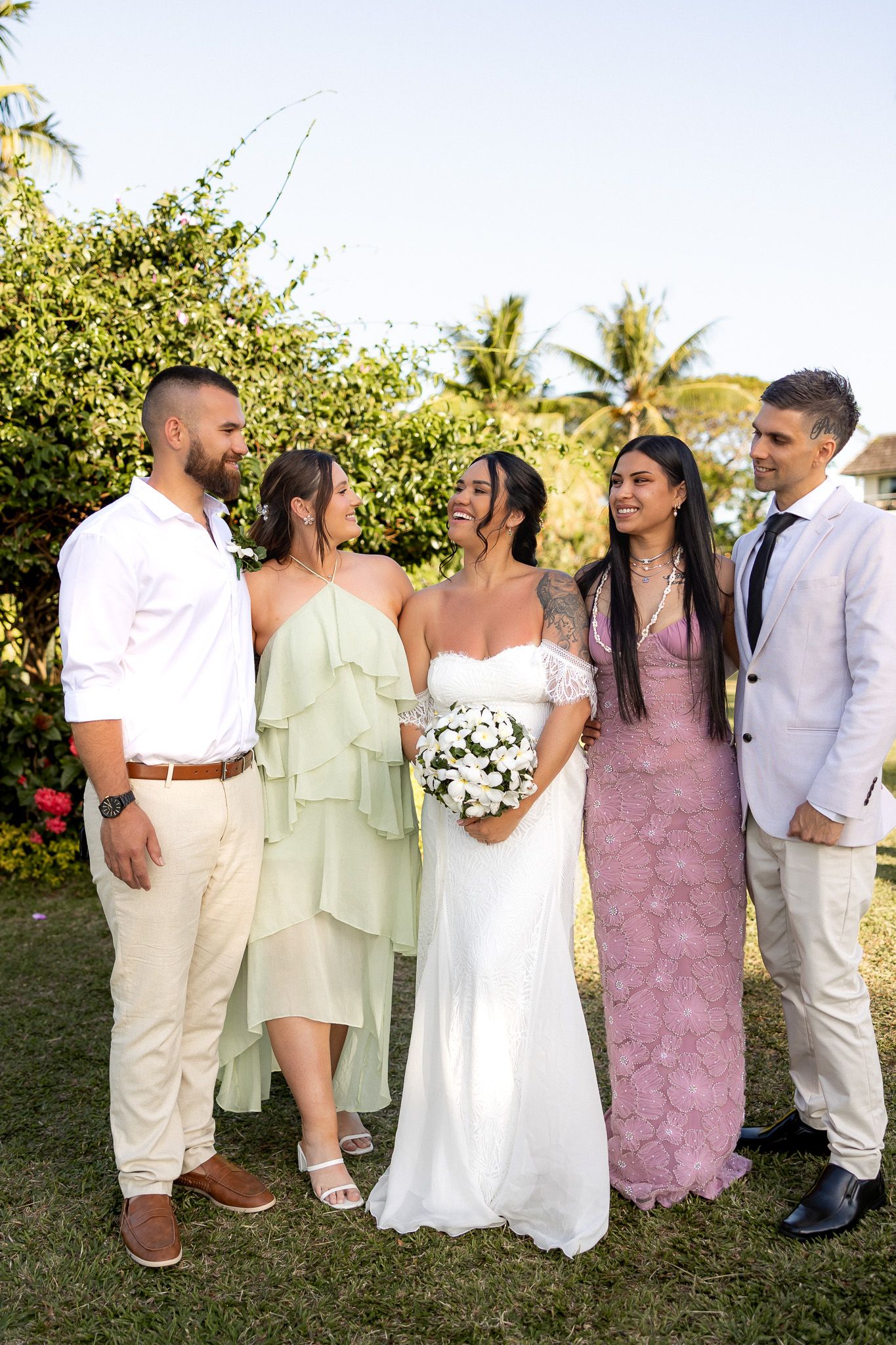 Bride with bouquet poses with four wedding guests in garden with tropical greenery backdrop.