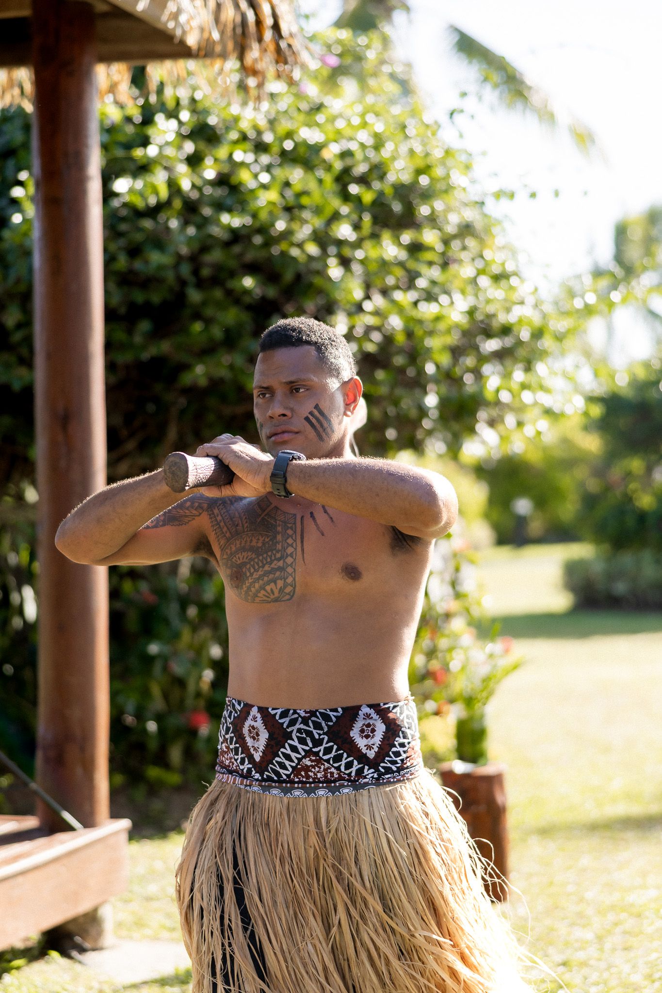 Shirtless man in traditional grass skirt and tribal tattoos performs cultural dance outdoors.