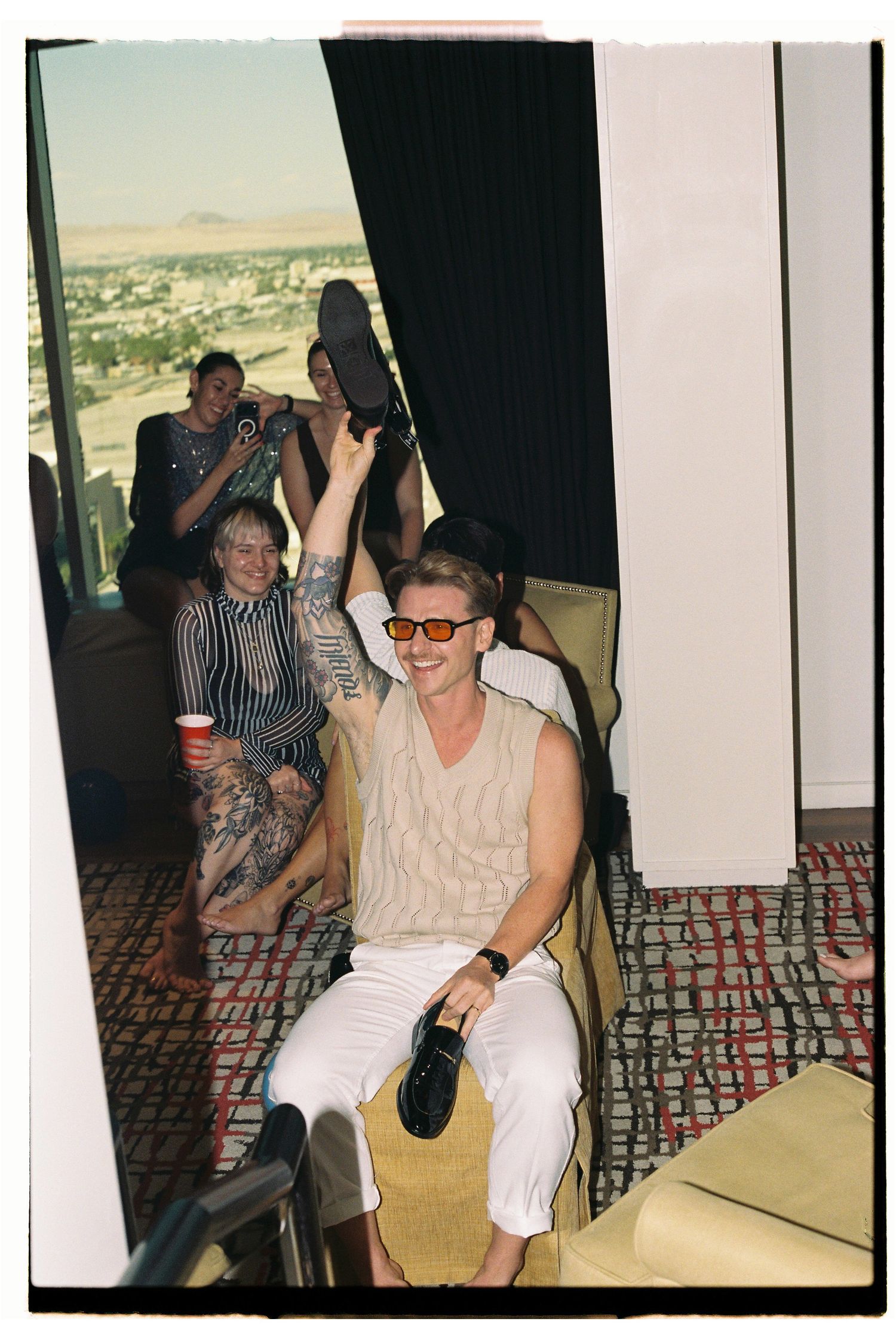 Man in sunglasses raises shoe at Las Vegas hotel party with friends gathered near window with city view.