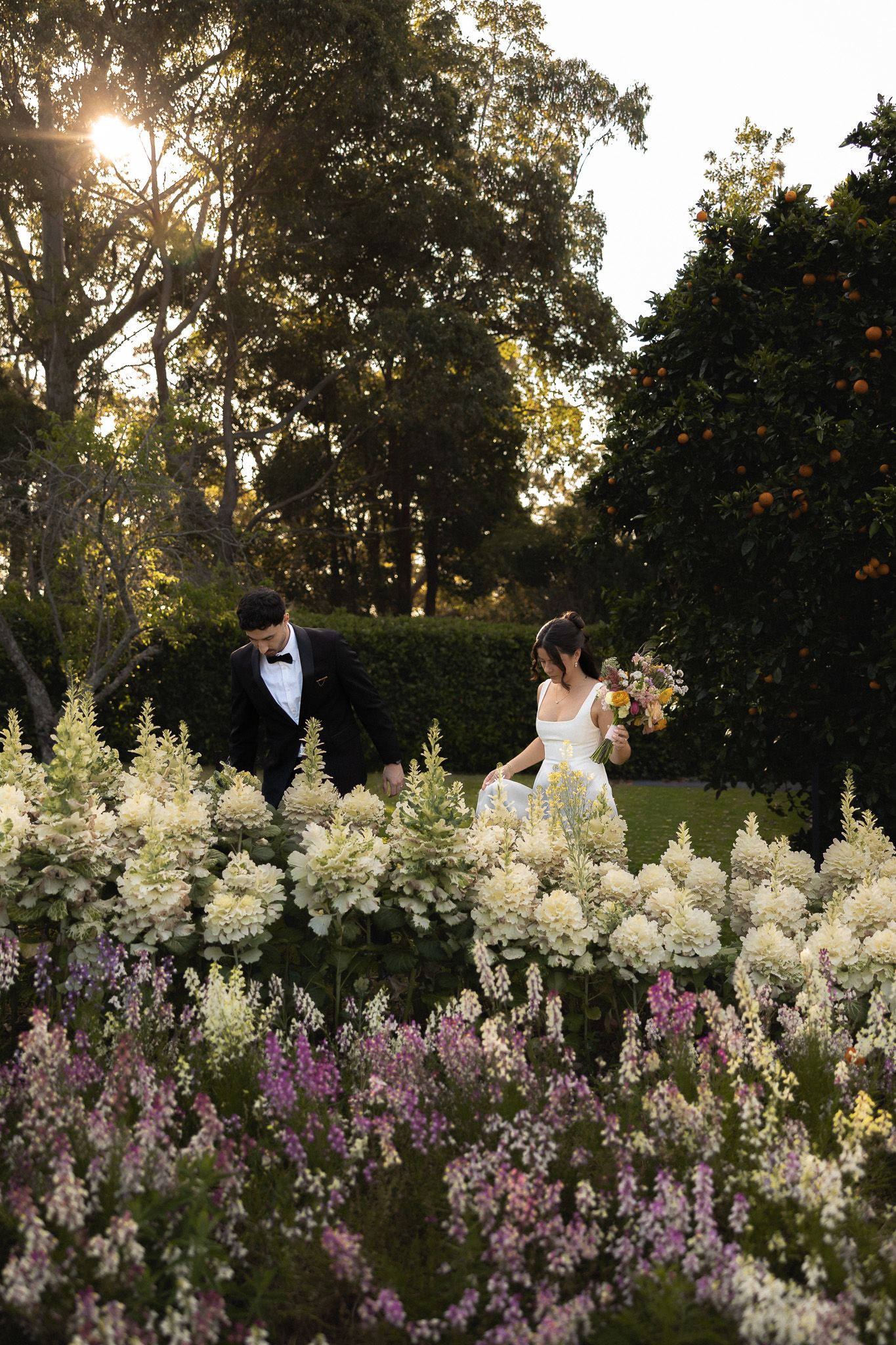 Bride and groom walking through lush garden with white and pink flowers at golden hour sunset.
