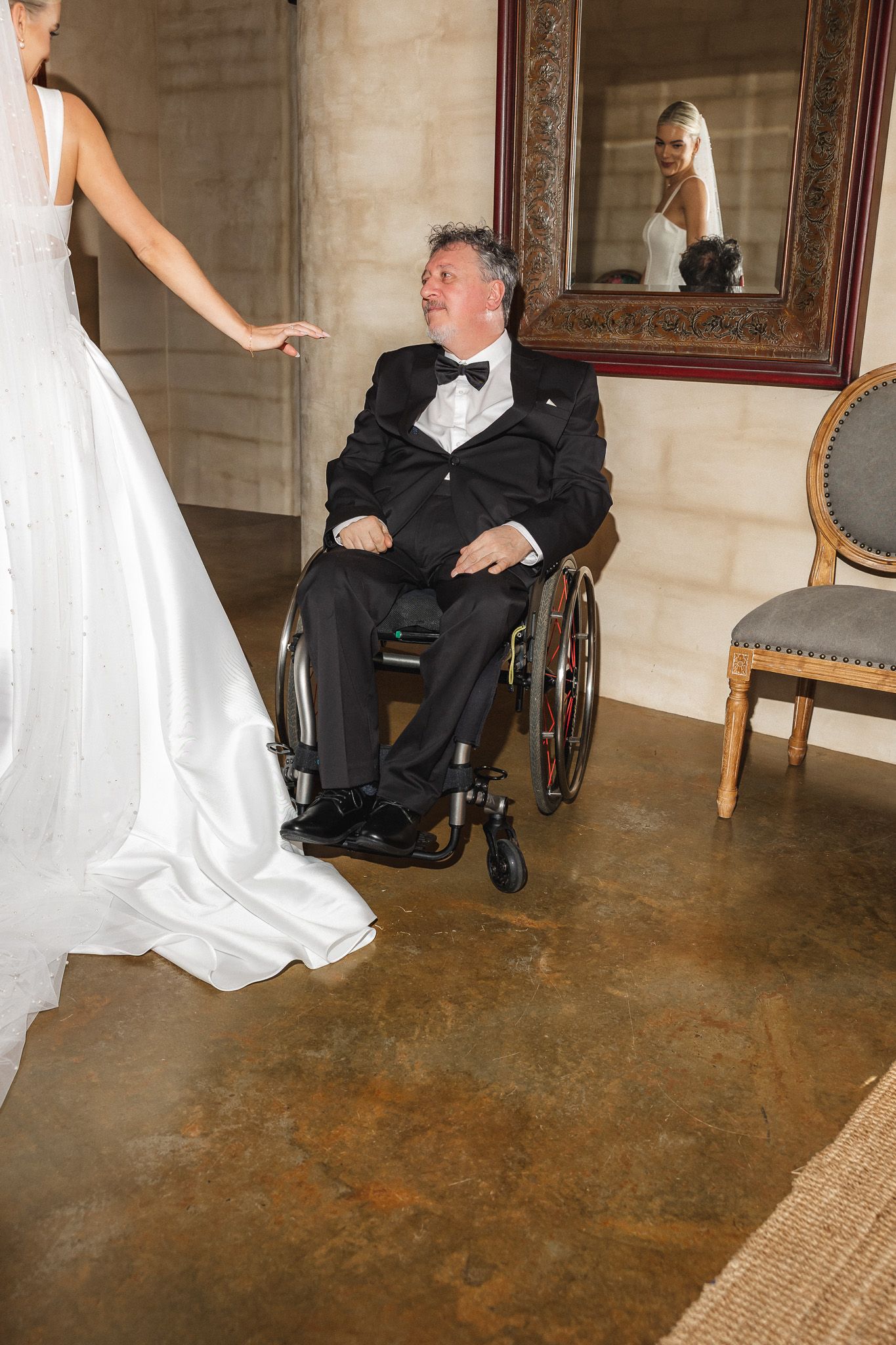 A bride in a white gown reaches toward an elderly man in a wheelchair wearing a tuxedo at a wedding venue.