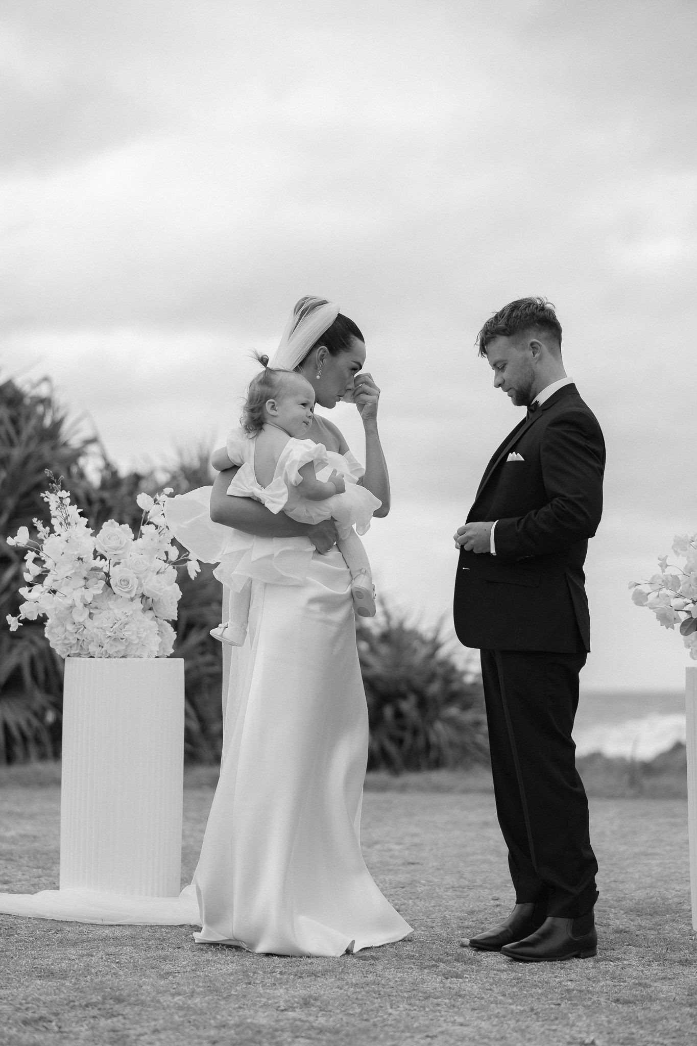 Black and white photo of bride holding baby while groom watches during outdoor wedding ceremony.