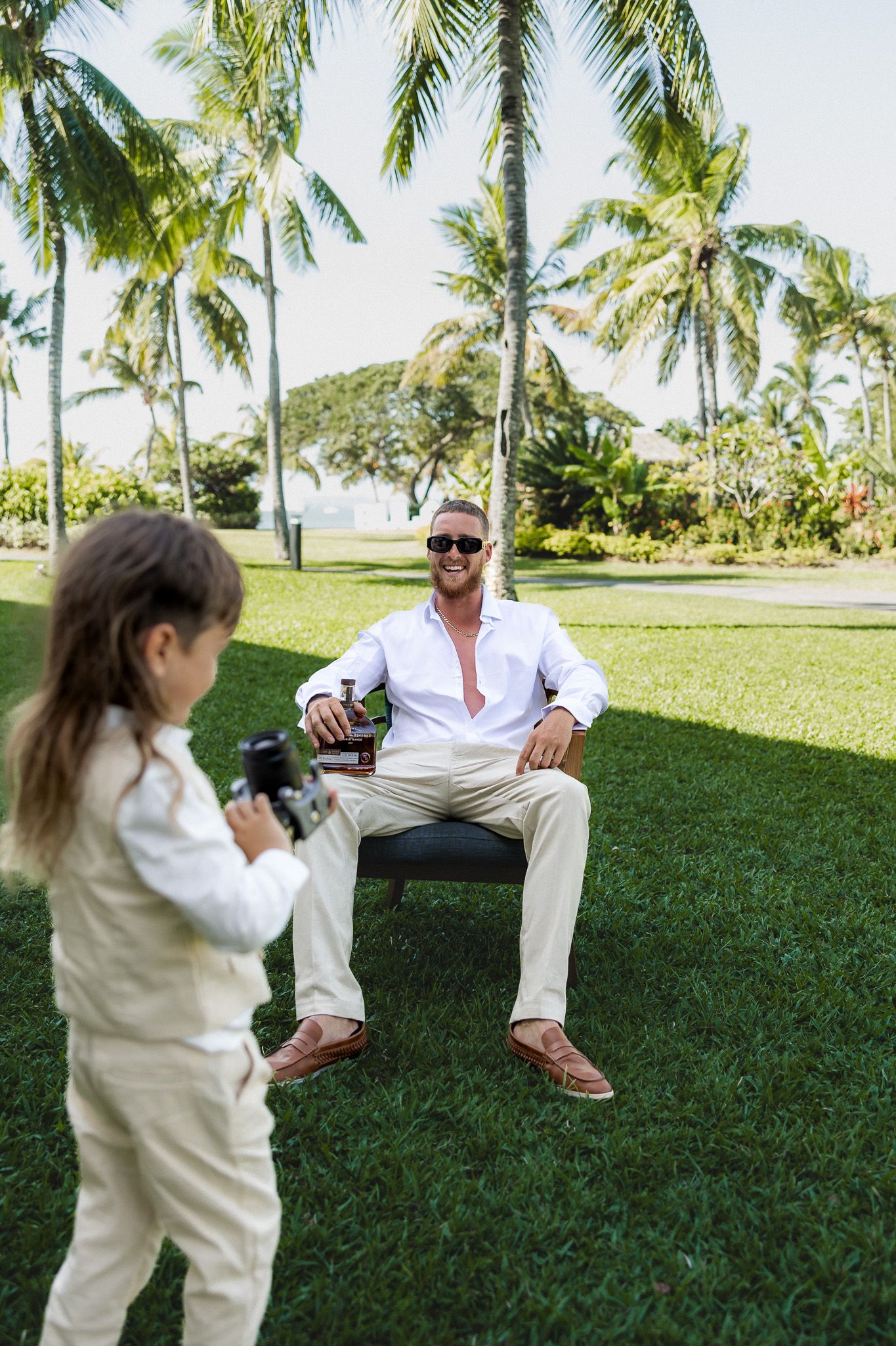 Man in white shirt and sunglasses poses with beer bottle while child photographs him in tropical garden.