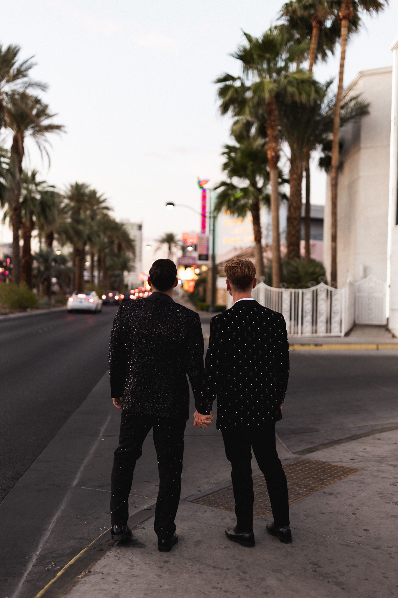 Two men in matching black suits hold hands walking along Las Vegas Strip lined with palm trees.