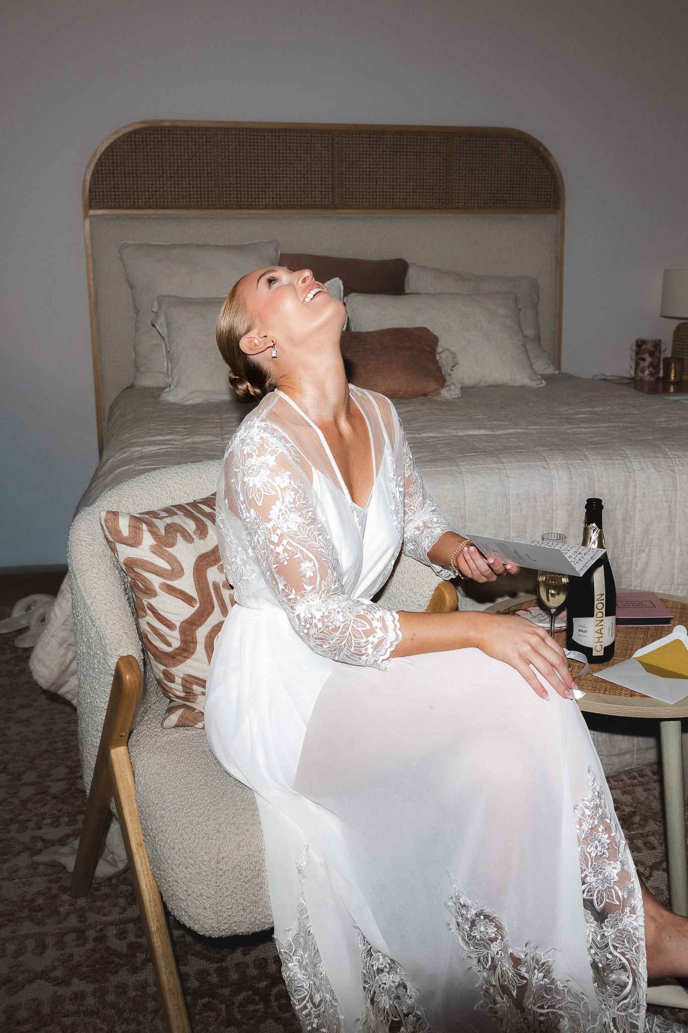 Happy bride in white lace robe laughing while seated in hotel room chair.