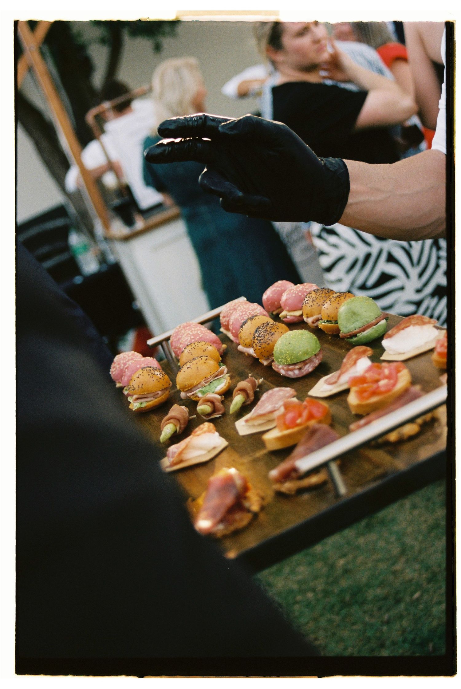 Caterer in black gloves arranging colorful sliders and appetizers on wooden serving board.