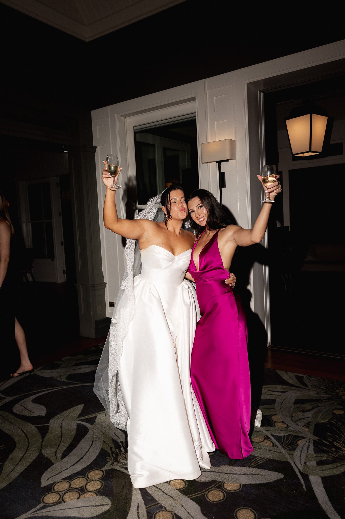 Bride in white strapless gown and guest in magenta dress celebrating with raised champagne glasses.