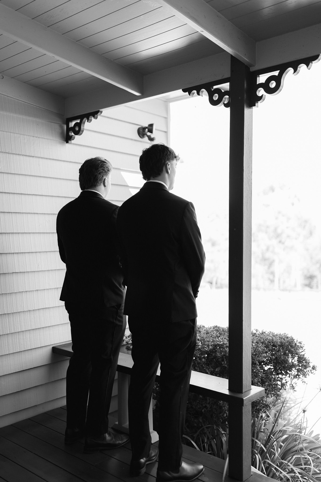 Two men in dark suits standing on a wooden porch viewed from behind in black and white.