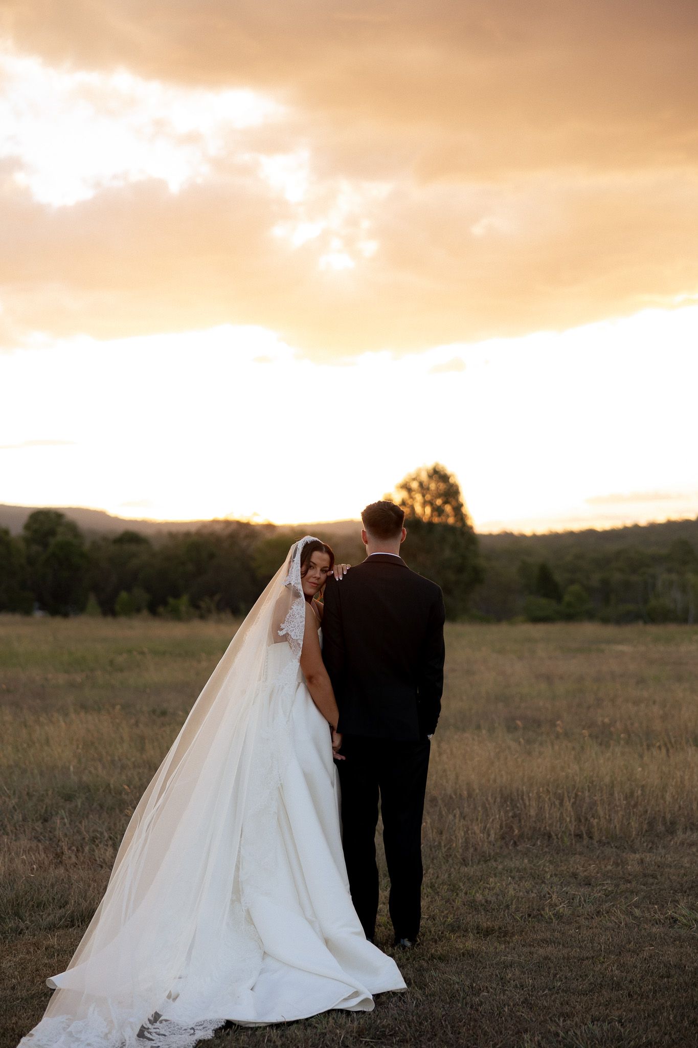 Bride and groom embracing in open field during golden sunset.