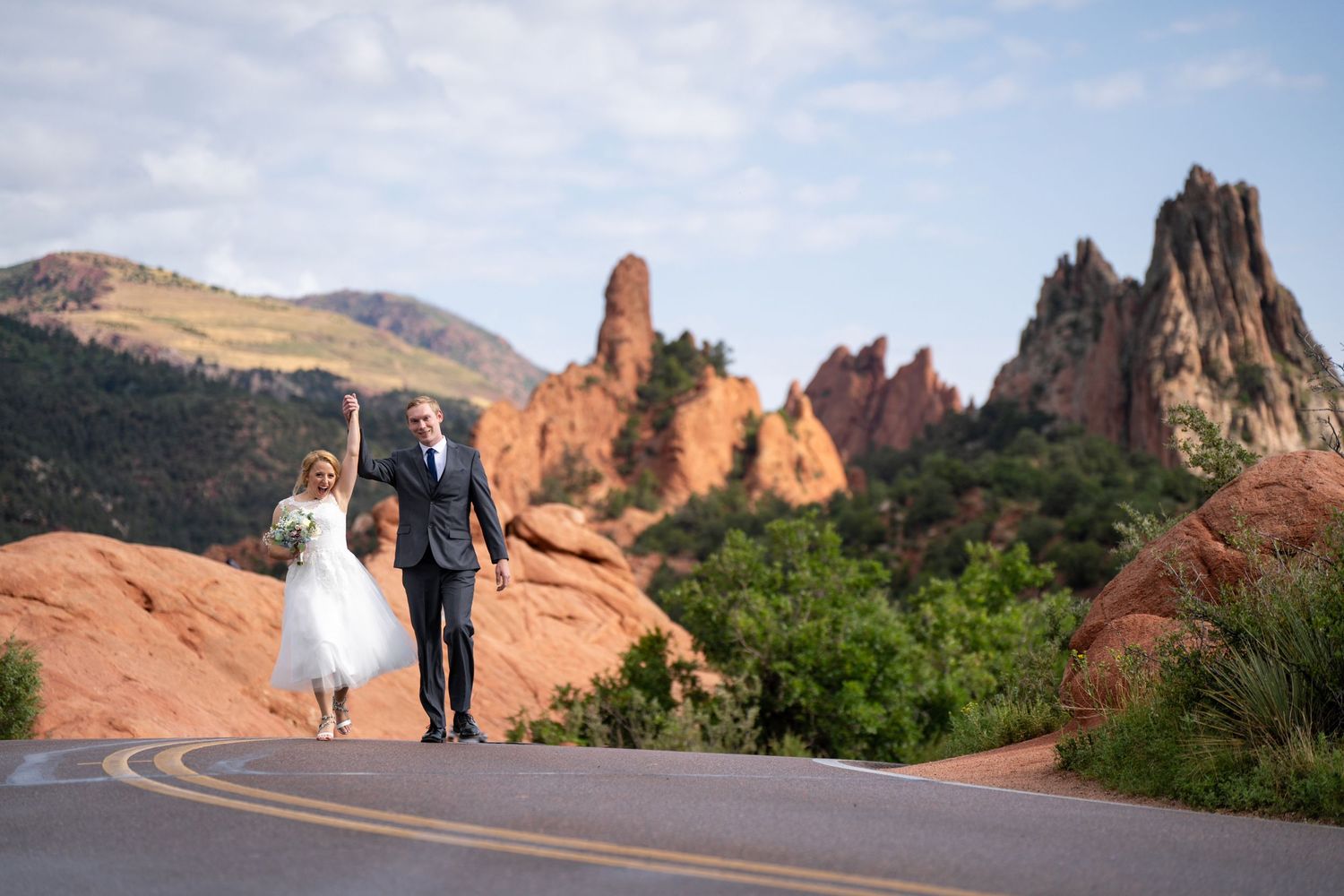 Bride and groom walk hand in hand down road with stunning Garden of the Gods red rocks backdrop.