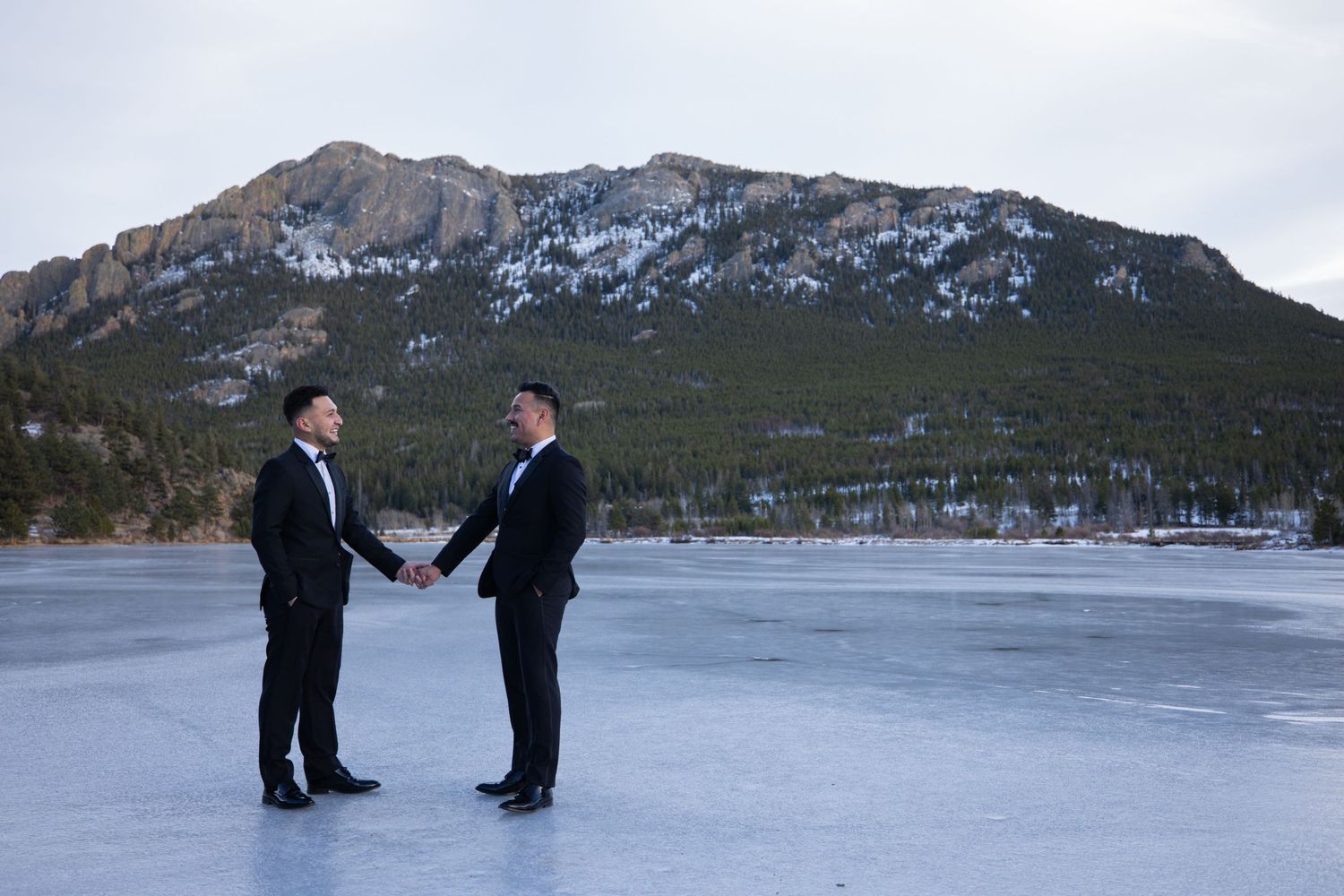 Two grooms in black tuxedos holding hands on a frozen lake with a mountain backdrop.