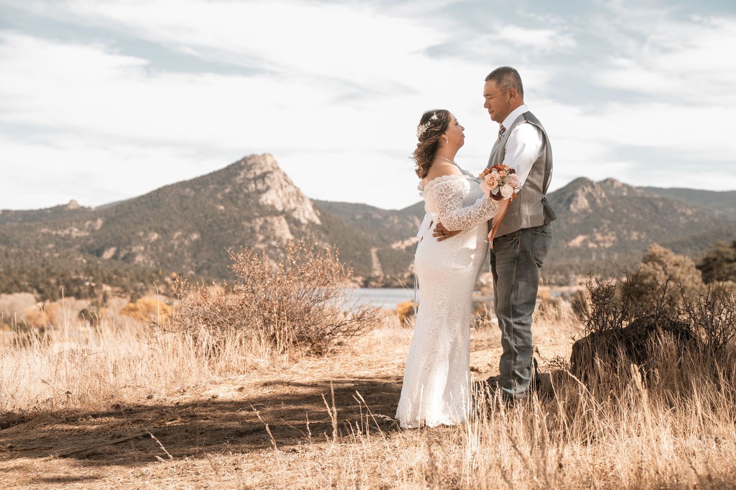 A bride and groom stand together in a dry meadow with rocky mountain peaks in the background.