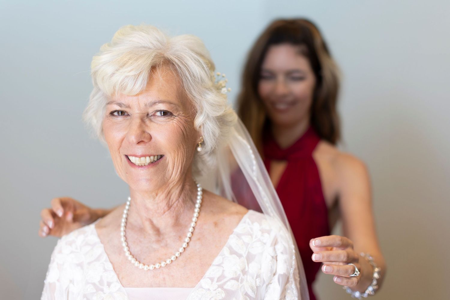Elderly bride in white lace dress and pearl necklace smiles while bridesmaid in red adjusts her veil.