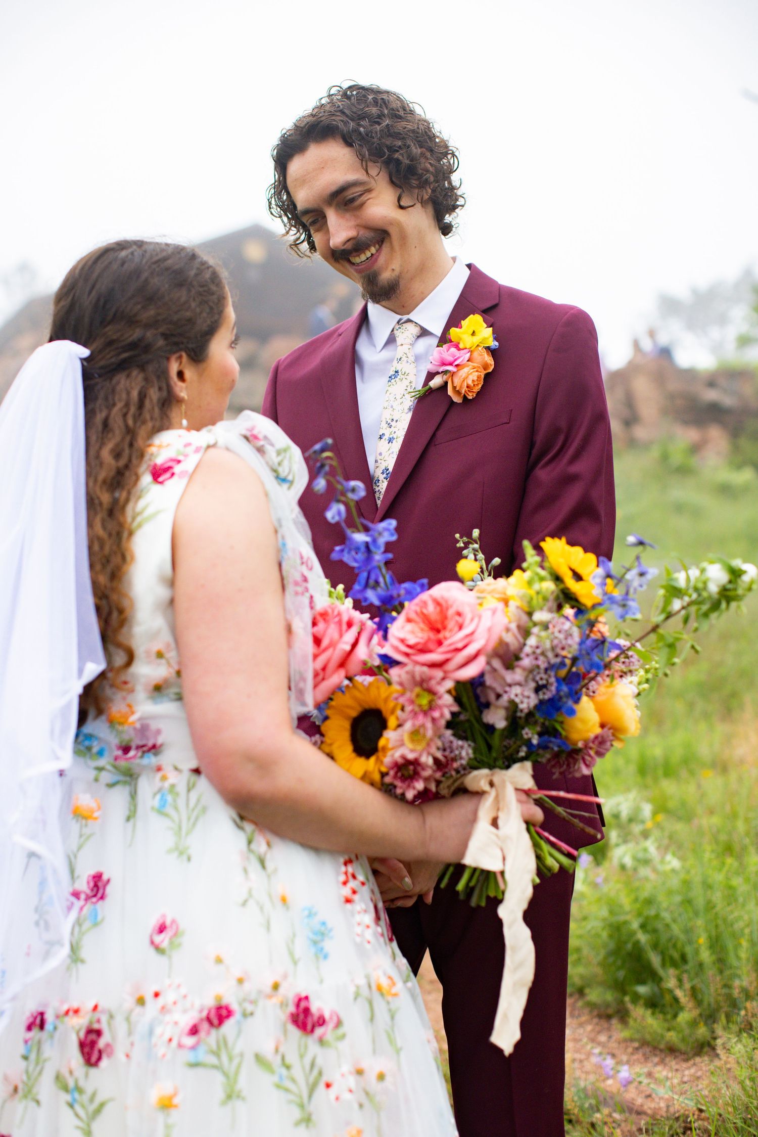 Smiling groom in burgundy suit holding hands with bride in floral dress holding vibrant bouquet outdoors.