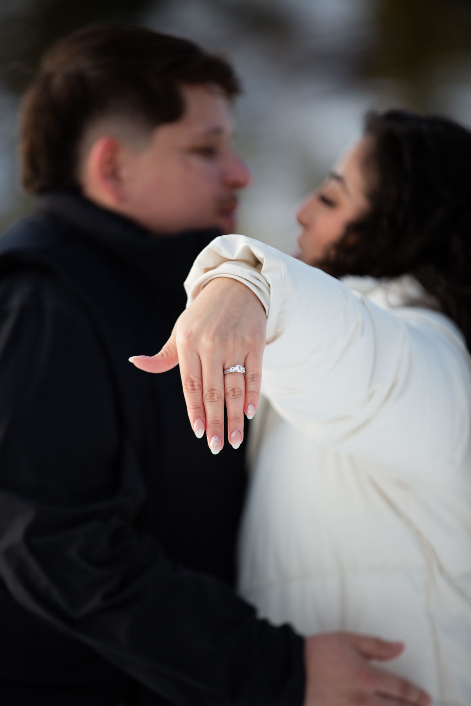 Engaged couple kissing in winter, woman in white coat extending hand to show diamond engagement ring.