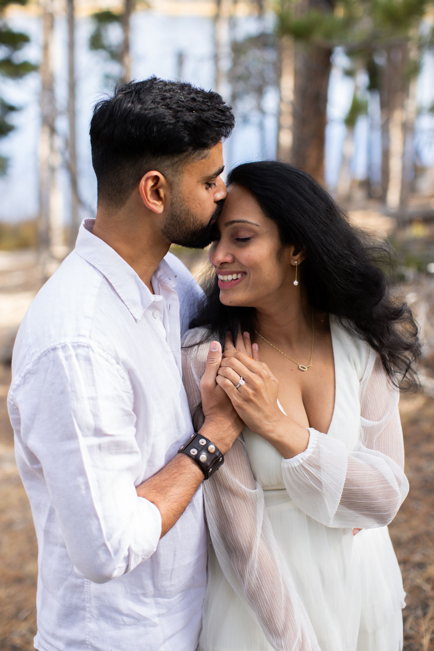 South Asian couple embraces among pine trees, man in white kisses womans forehead as she smiles.