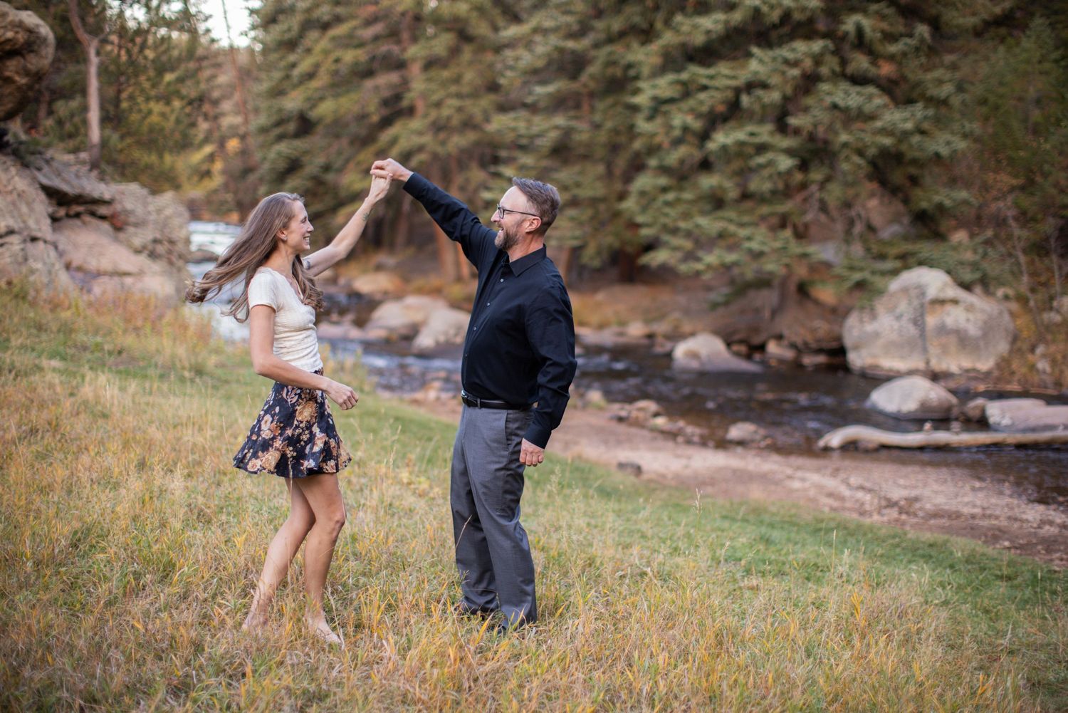 Couple dancing in a meadow near a stream, man spinning woman in floral skirt among autumn grasses.