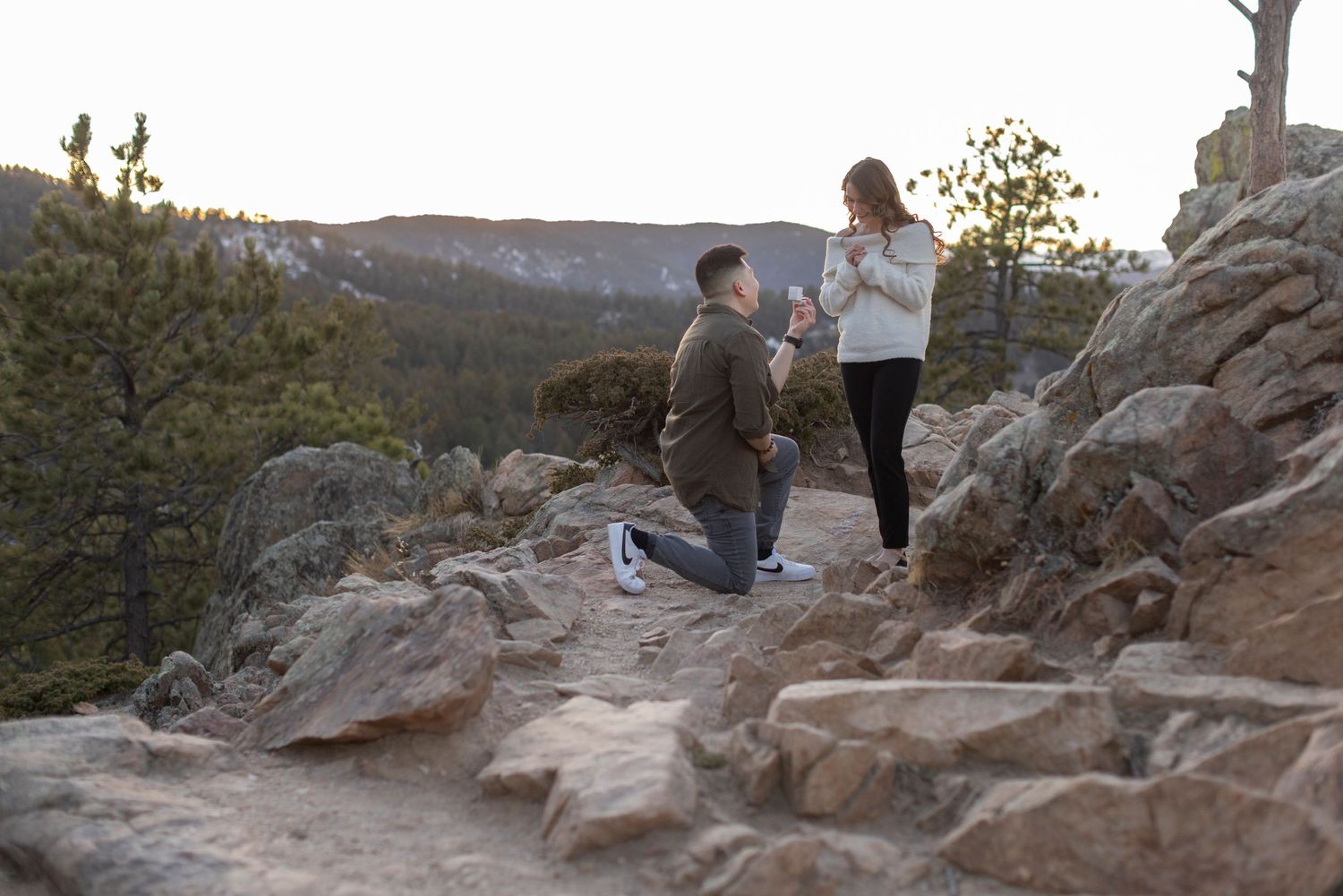 Man proposes to woman on rocky mountain overlook at sunset surrounded by pine trees and scenic landscape.