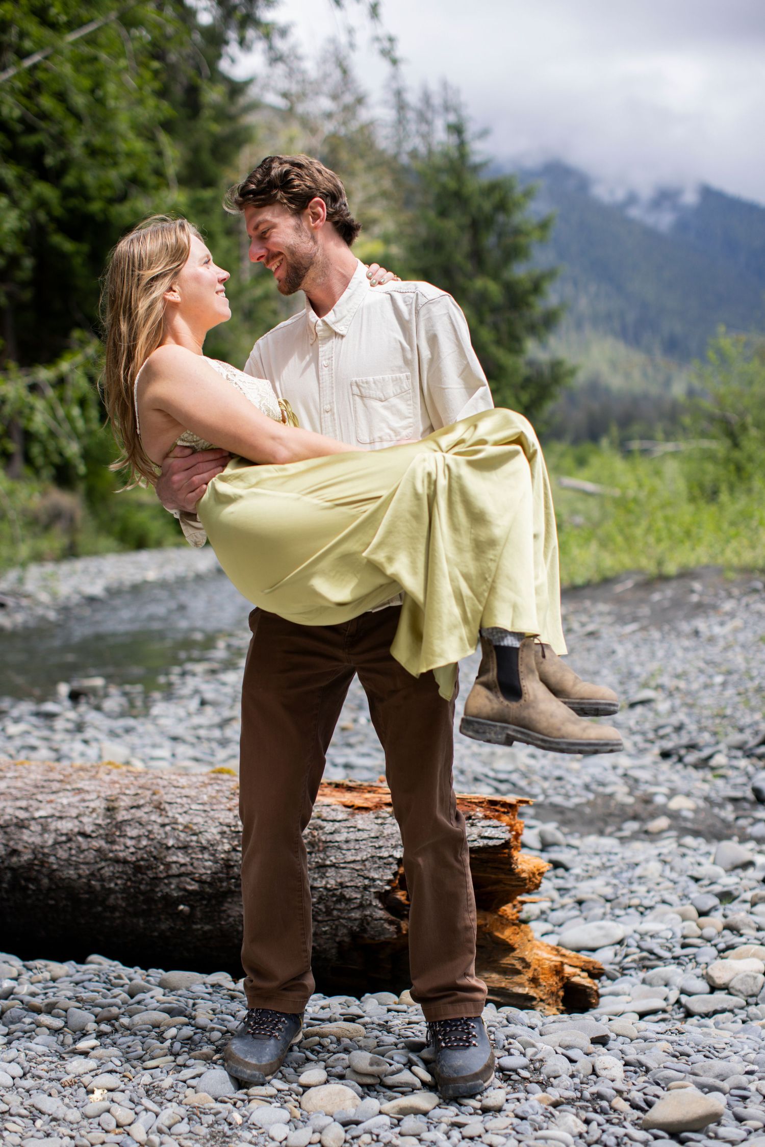 A smiling man carries a woman in a yellow dress along a rocky mountain riverbank.