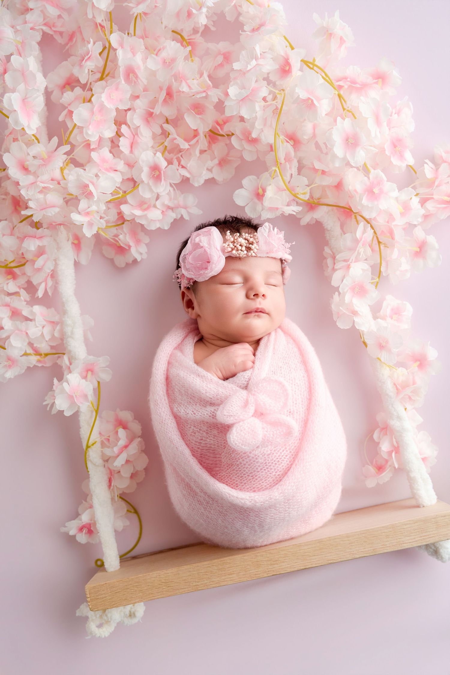 Newborn baby girl swaddled in pink wrap sleeping on a wooden swing surrounded by cherry blossom flowers.