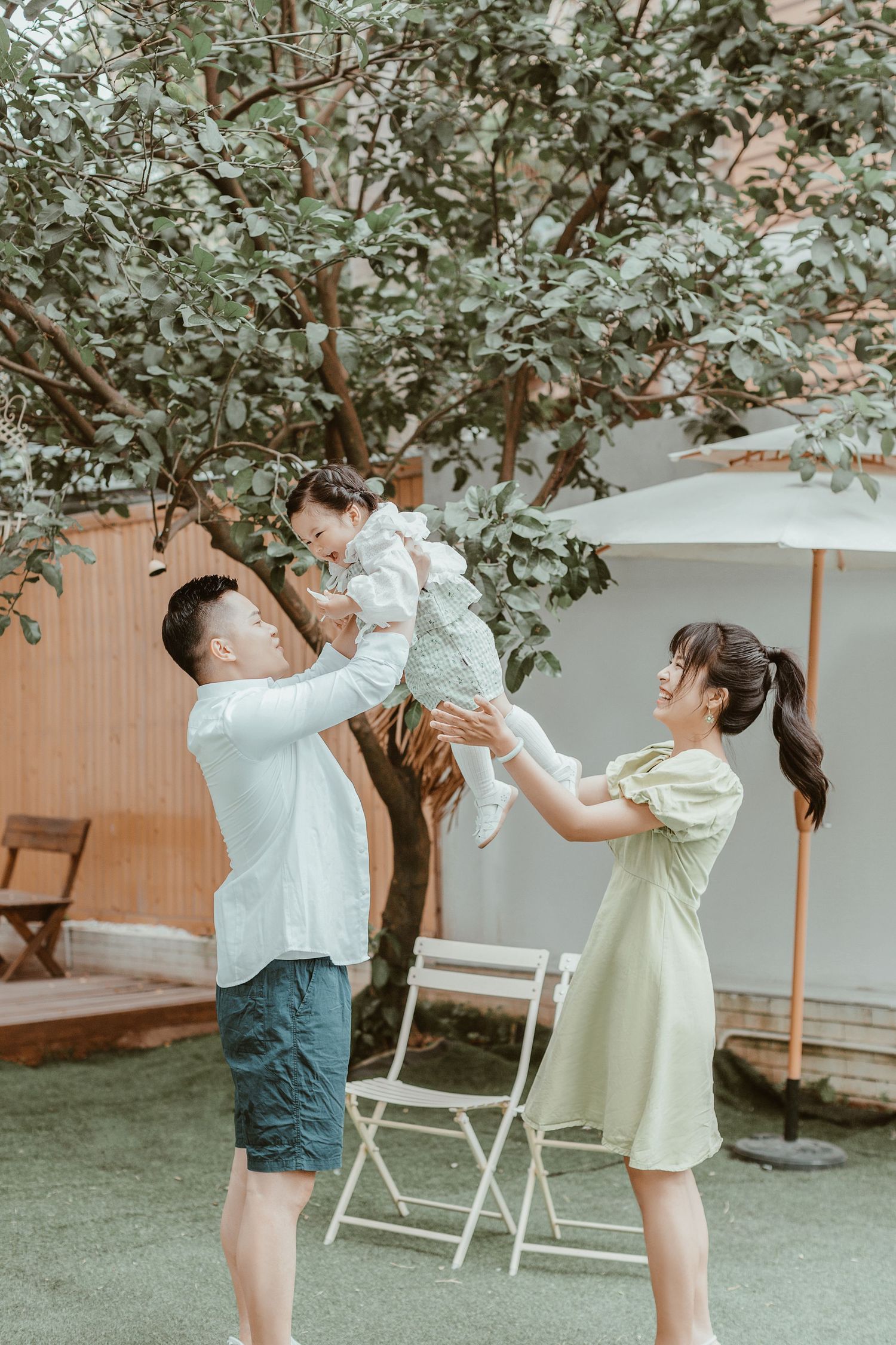 Joyful Asian parents lift their laughing toddler high in the air in a beautiful garden setting.