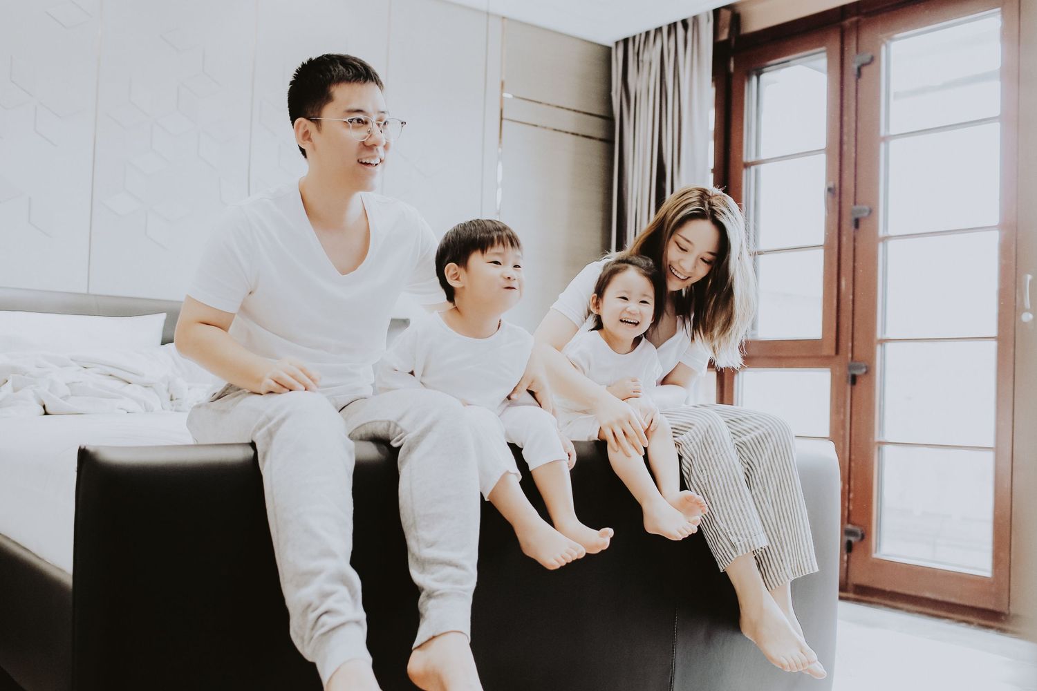 An Asian family of four sits casually on a dark couch near a bright window, smiling and relaxed.
