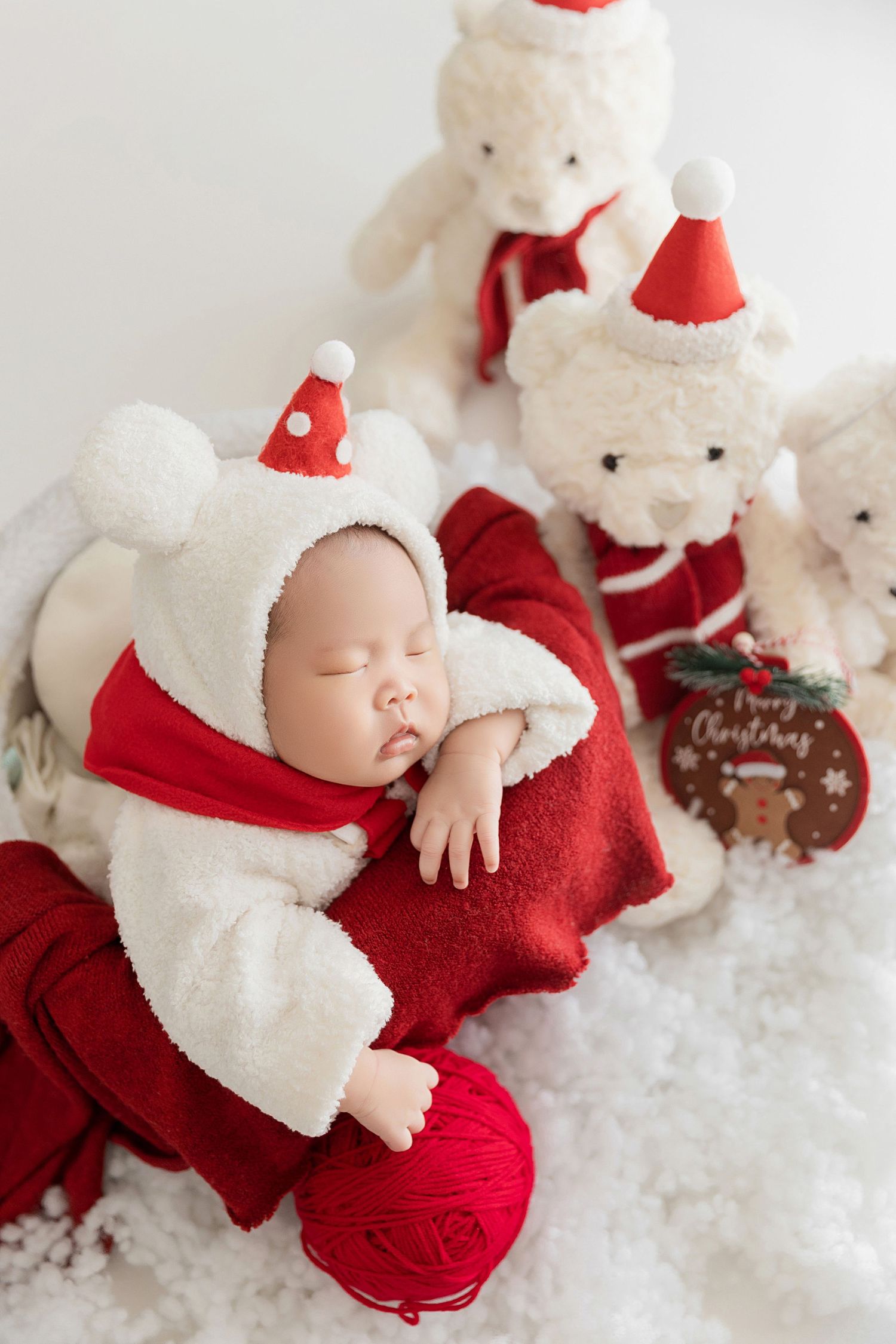 Newborn baby in white Santa costume sleeping beside white Christmas teddy bears on snowy white background.