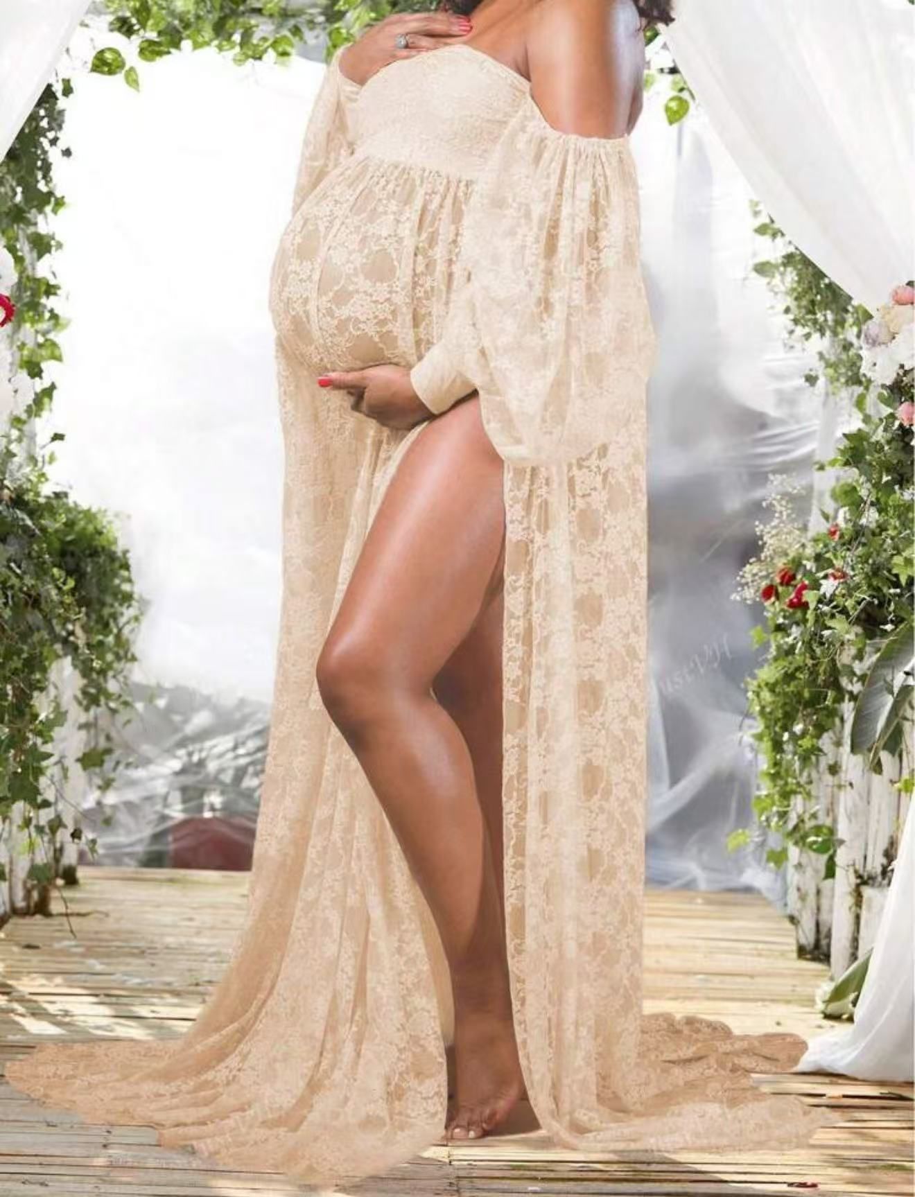 Pregnant woman in sheer champagne lace open-front maternity gown posing under floral arch outdoors.
