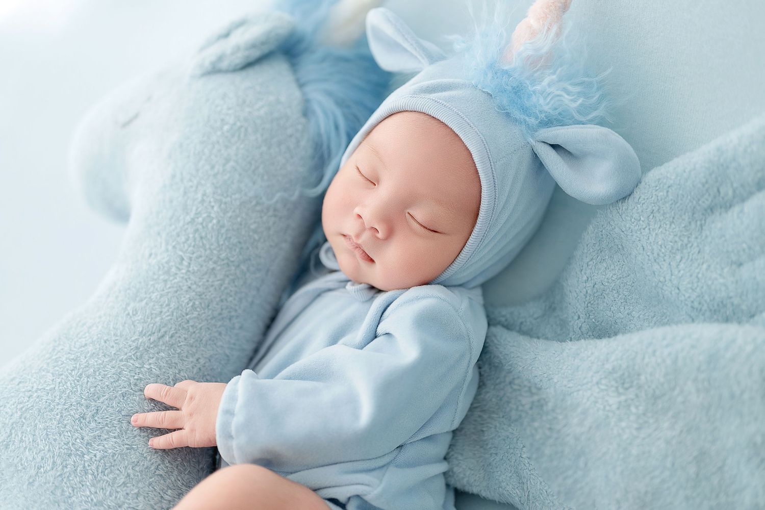 Sleeping newborn baby in blue unicorn outfit and bonnet resting on soft blue blanket with plush toy.