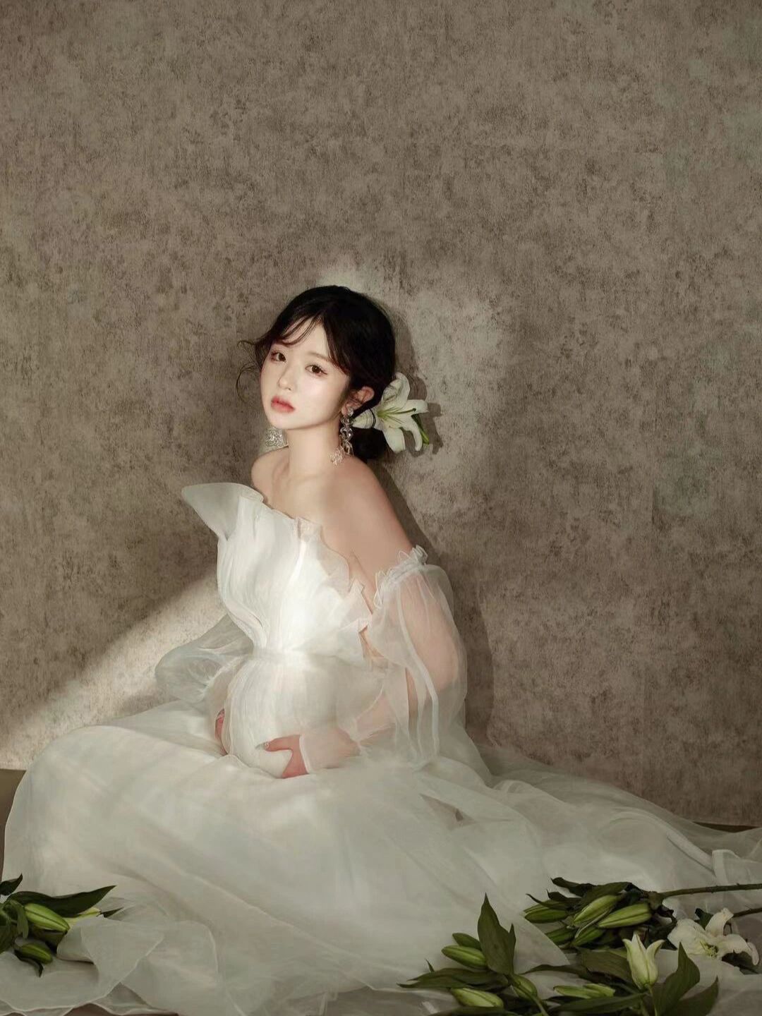 Woman in white tulle maternity gown sitting gracefully among white lilies in elegant pregnancy portrait.