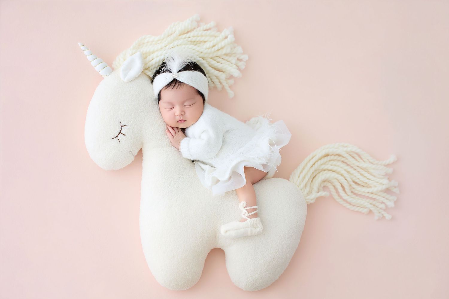 Newborn baby sleeping in white outfit on a white unicorn stuffed animal against a soft pink background.