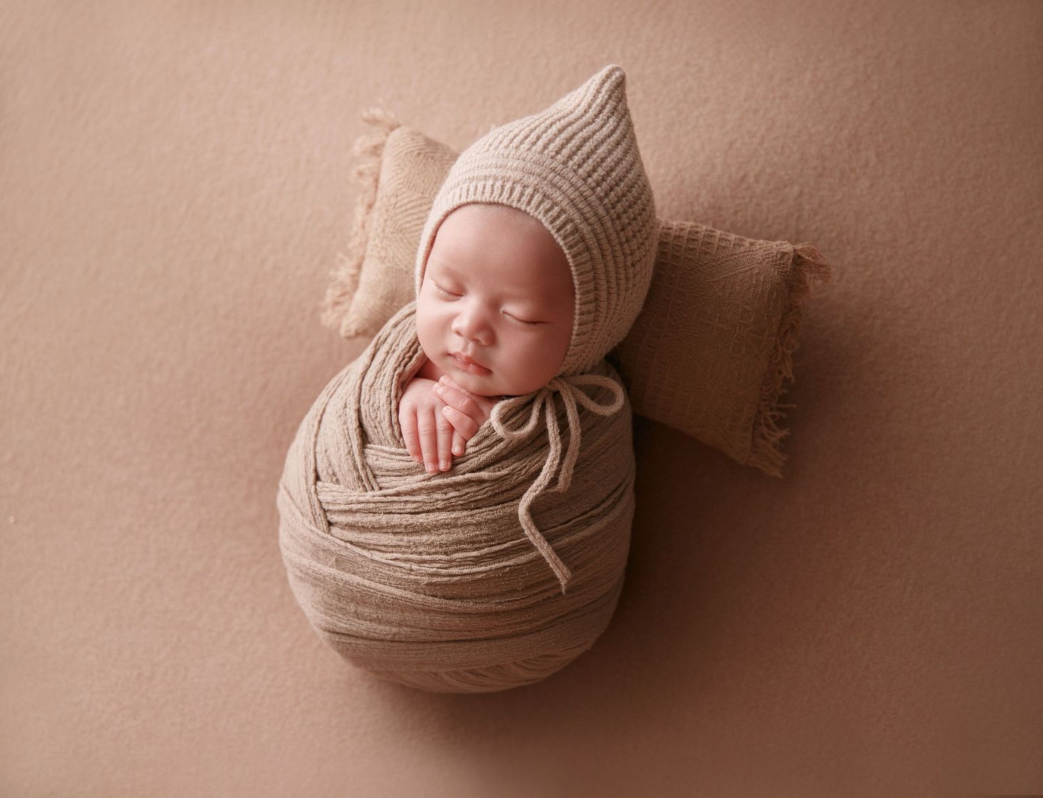 Sleeping newborn baby snugly wrapped in beige knit swaddle wearing pointed bonnet on small pillow.