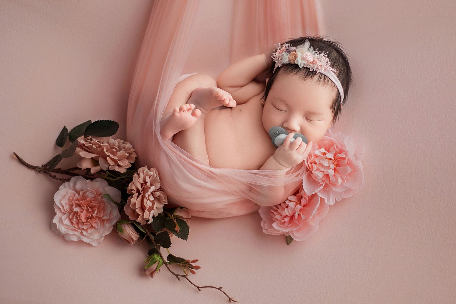 Peaceful newborn baby sleeping on pink tulle fabric with blush peonies and delicate floral headband.