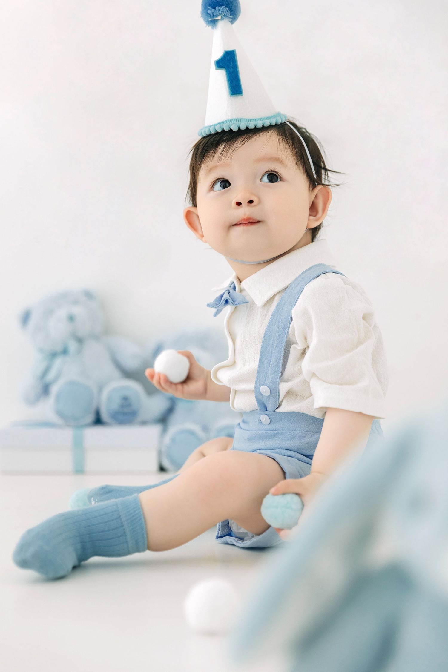 Adorable baby boy in blue suspenders and birthday hat celebrating his 1st birthday with stuffed animals.