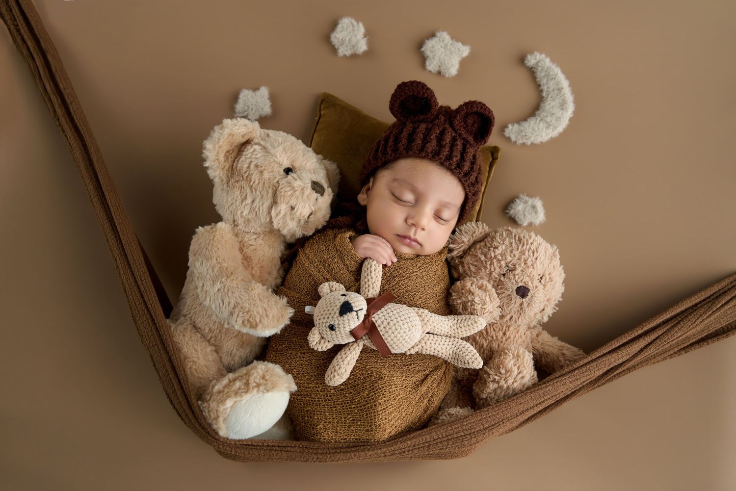 Newborn baby sleeping in hammock with teddy bears, wearing brown bear hat, surrounded by stars and moon decorations.