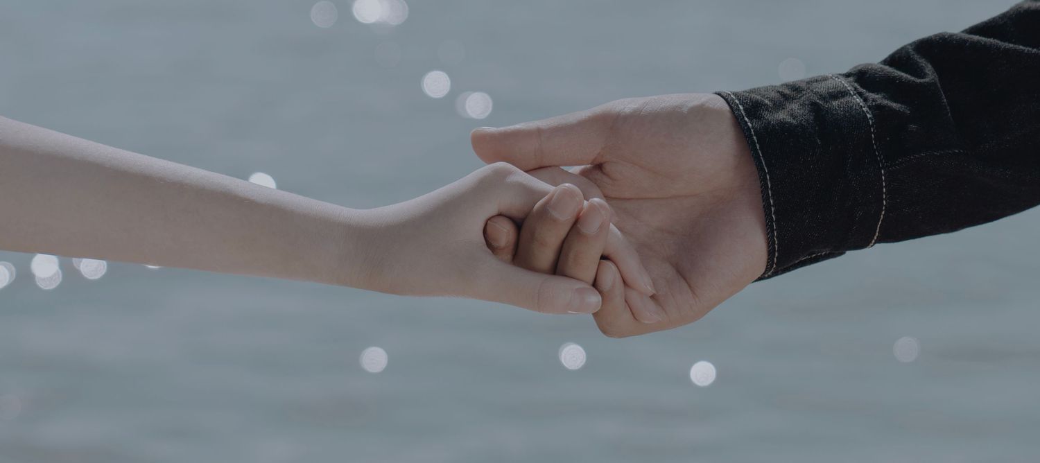 Two people gently holding hands near a shimmering water backdrop in soft natural light.