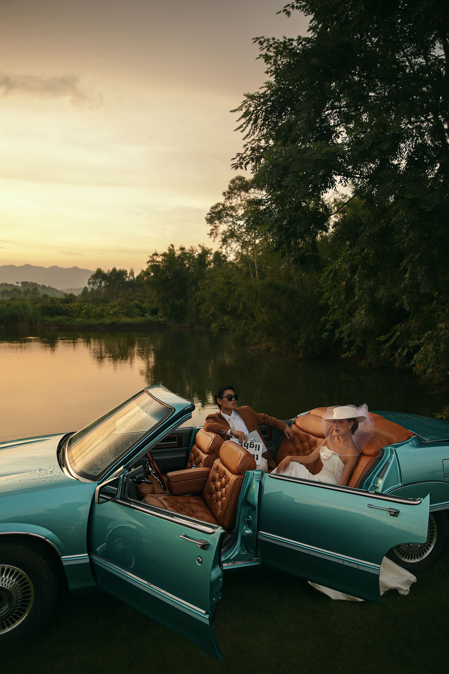 Bride and groom sitting in a teal vintage convertible convertible by a river at golden sunset.